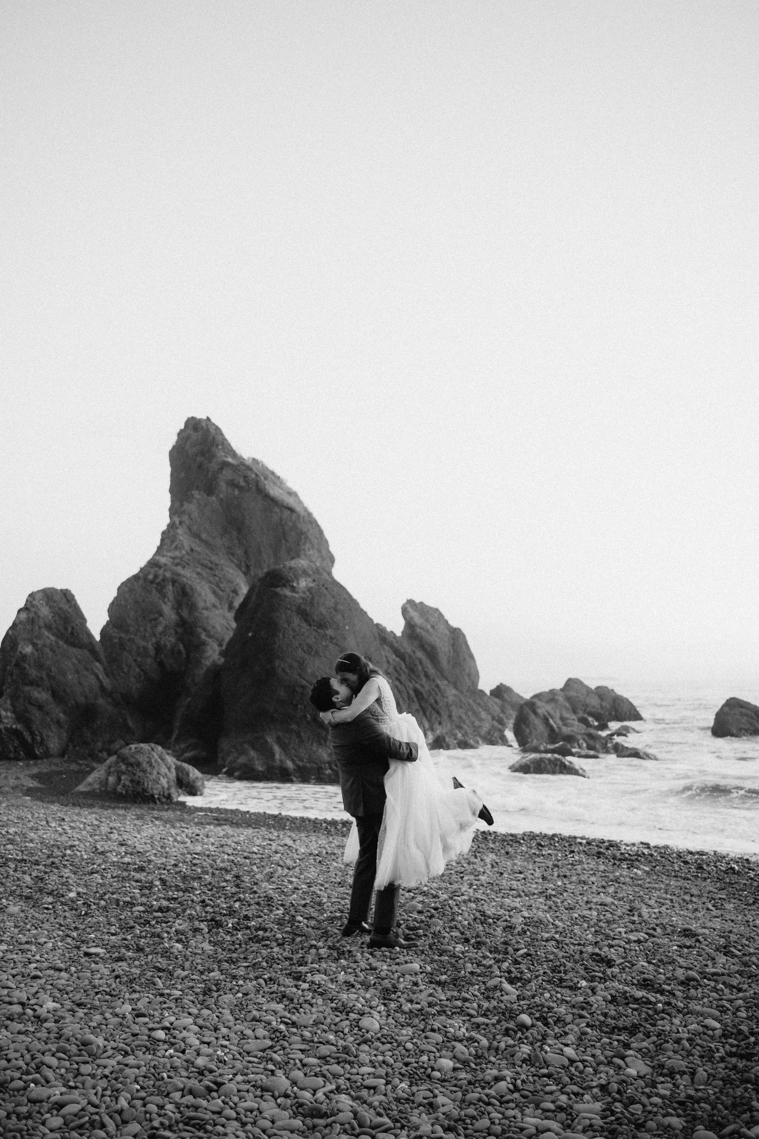 A black and white wedding photo of a couple dancing by dramatic coastal rock formations.
