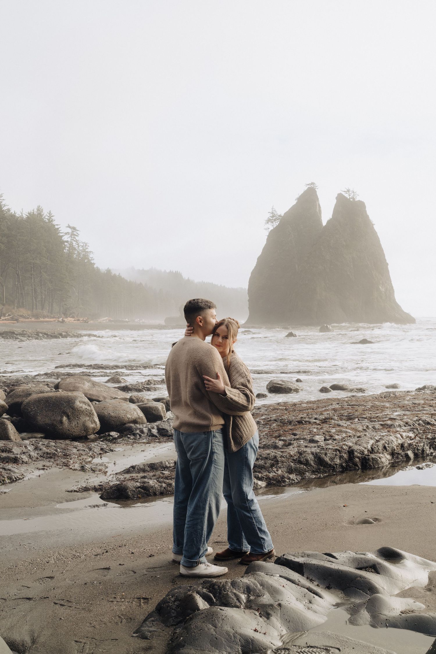 Two people embrace on rocky Pacific Northwest beach with sea stacks and misty coastline in the background.