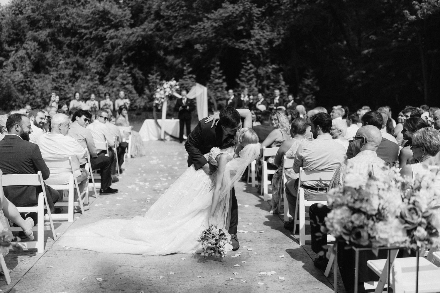 Outdoor wedding ceremony with guests seated in white chairs surrounded by lush greenery.
