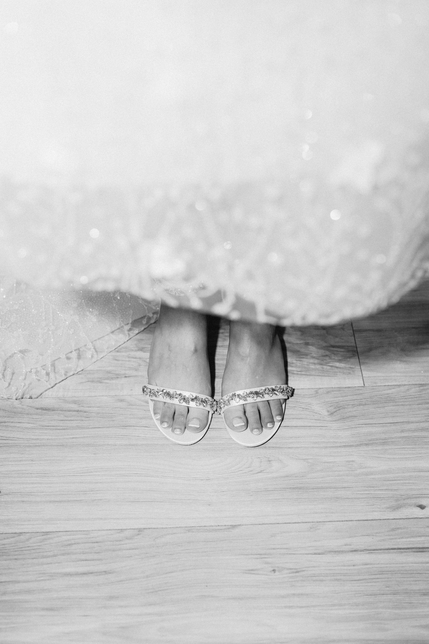 Close-up black and white photo of bare feet peeking out from under a white dress on wooden flooring.
