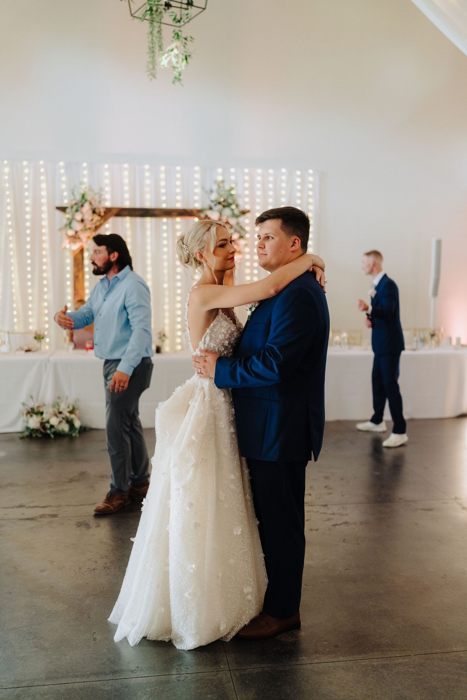 A wedding couple shares their first dance in a modern white venue decorated with string lights and floral arrangements.