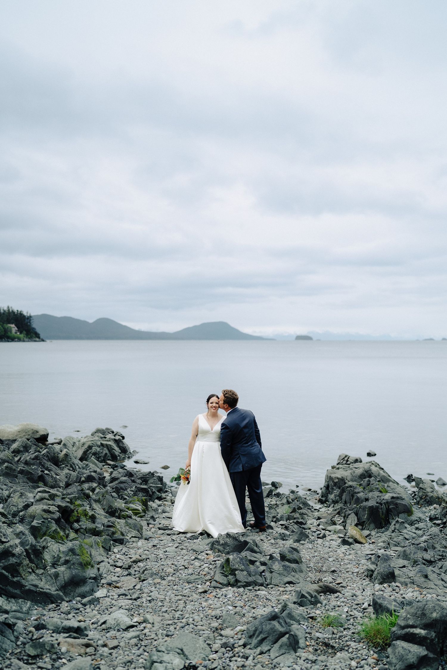 A couple in wedding attire poses on rocky coastal terrain with misty mountains in the background.