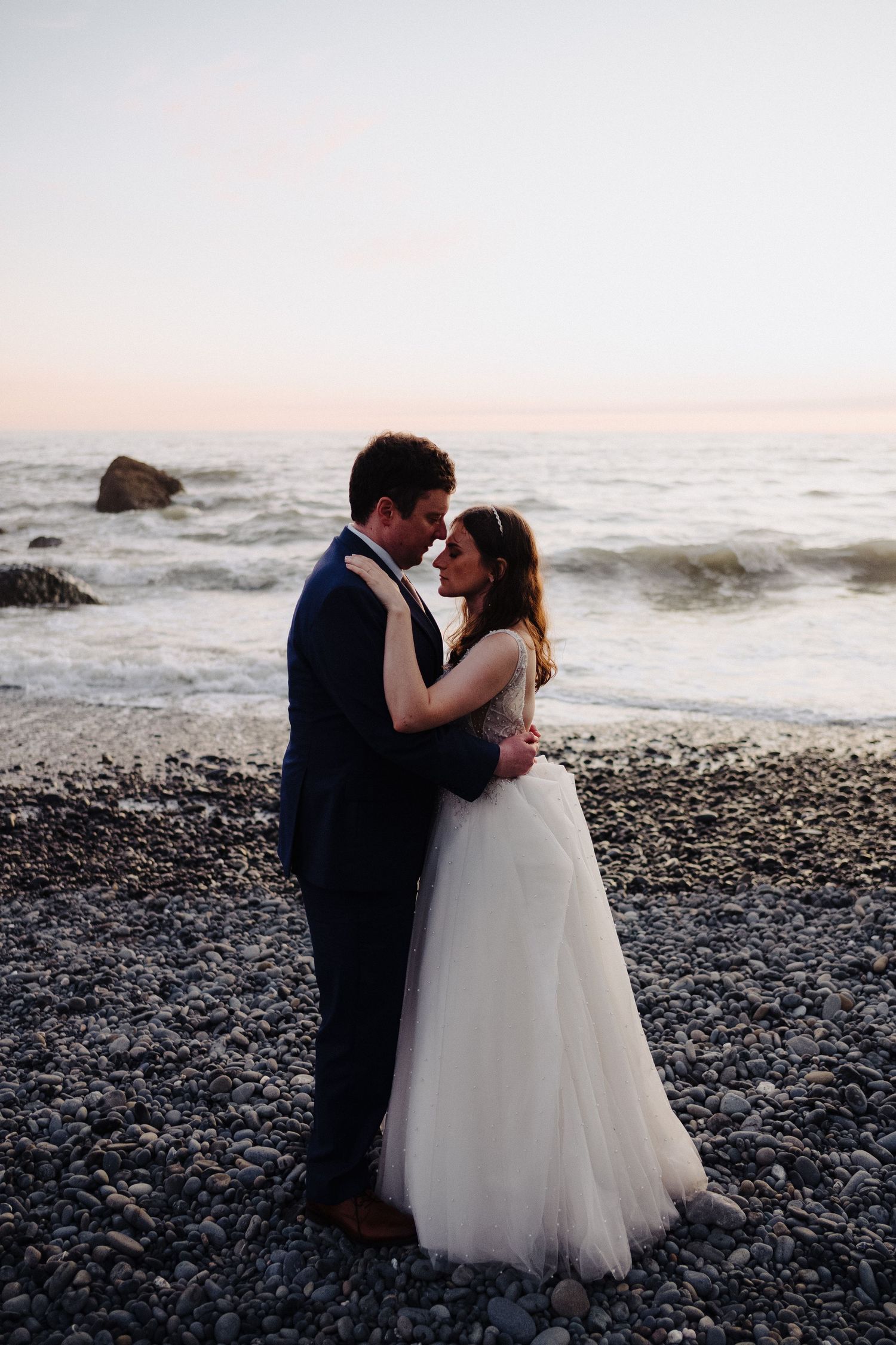 Romantic sunset beach portrait of newlyweds embracing on rocky shoreline.
