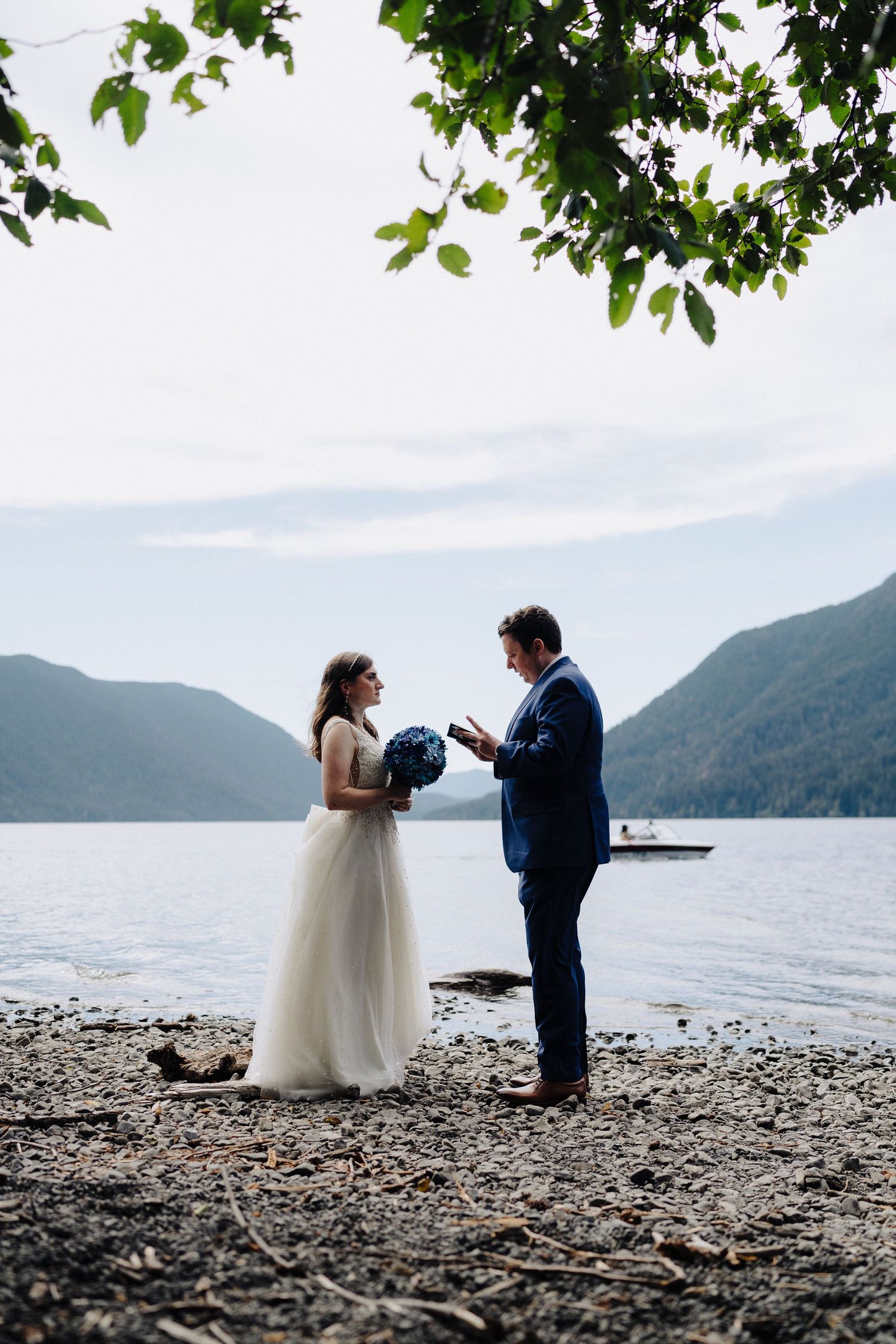 A wedding couple exchanges vows on a rocky beach shore with mountains and a lake in the background.