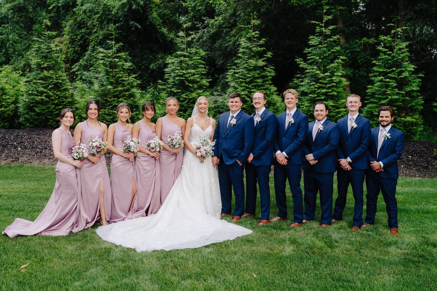 Wedding party in navy suits and blush dresses posed on green lawn.