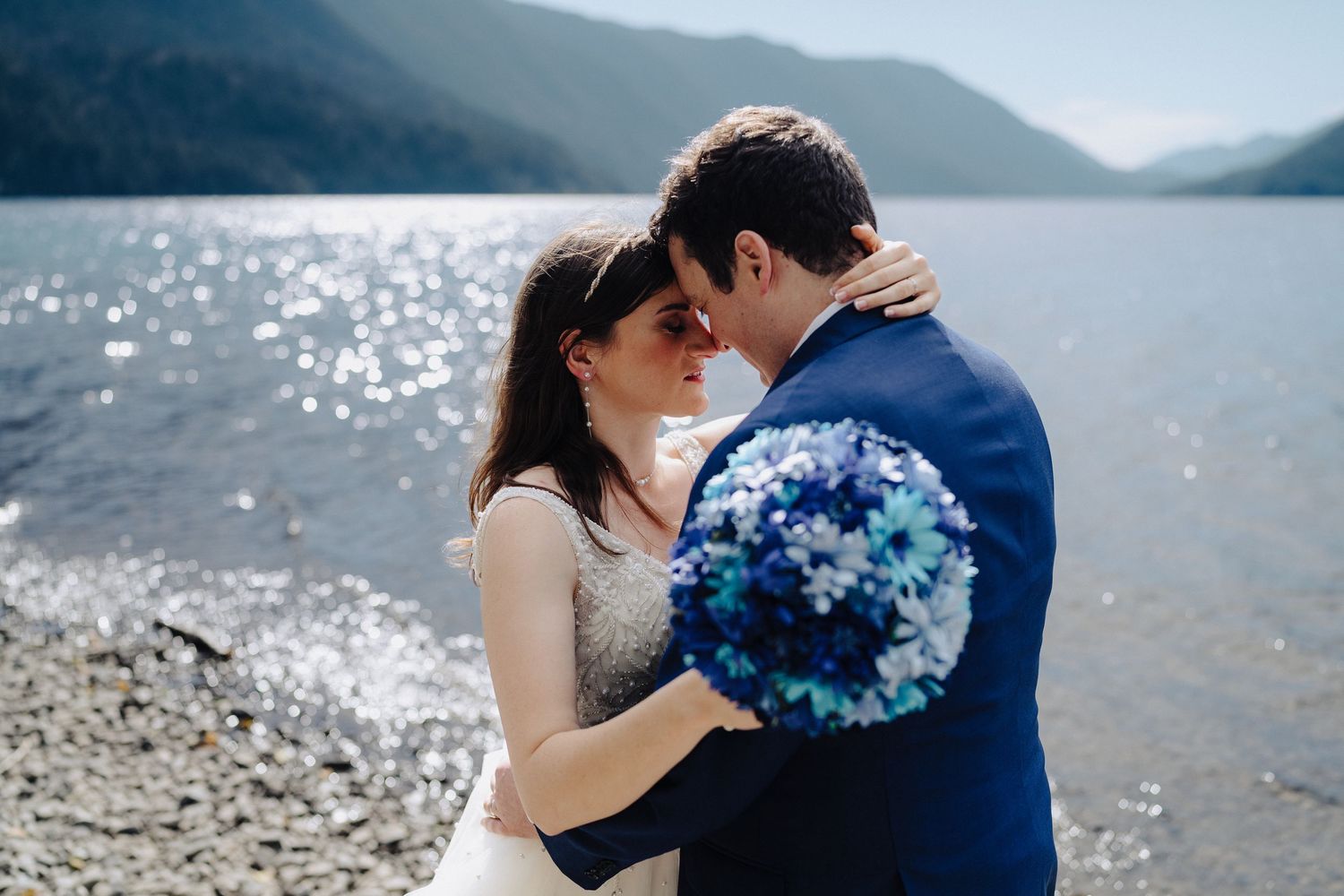 A couple shares an intimate moment on a lakeside beach while holding a blue bouquet.