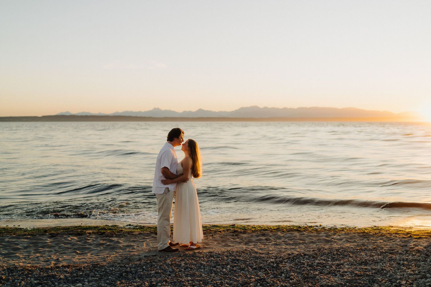 Romantic beach sunset scene with couple sitting by calm ocean waters during golden hour.