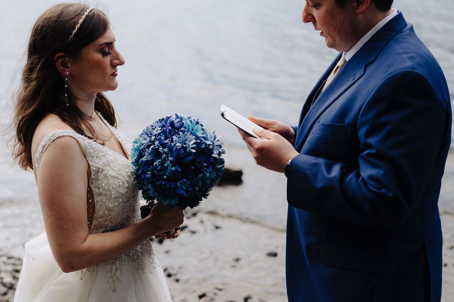 A wedding ceremony with vow reading takes place on a rocky lakeside beach.