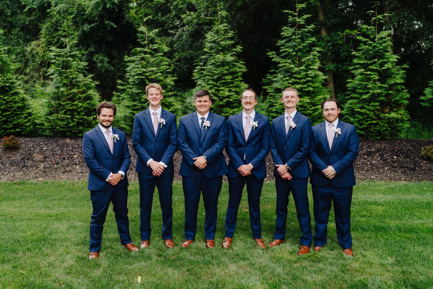Groomsmen in matching navy suits and brown shoes pose on green lawn for formal wedding photo.