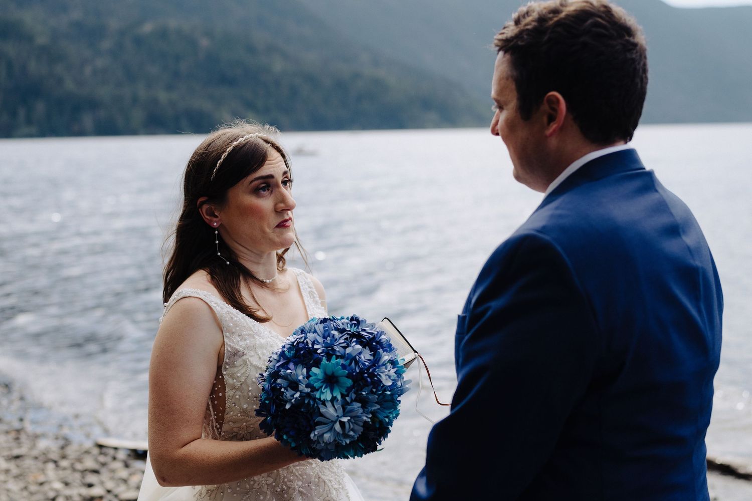 A bride holds a vibrant blue bouquet while standing on a lakeside beach surrounded by mountains.