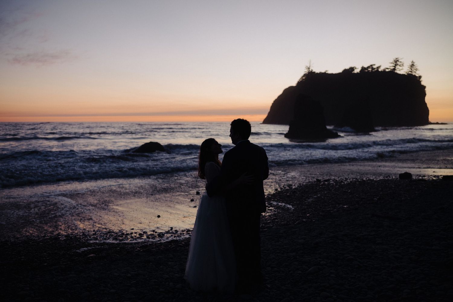 A silhouetted couple stands on a beach against a sunset sky with a sea stack in the background.