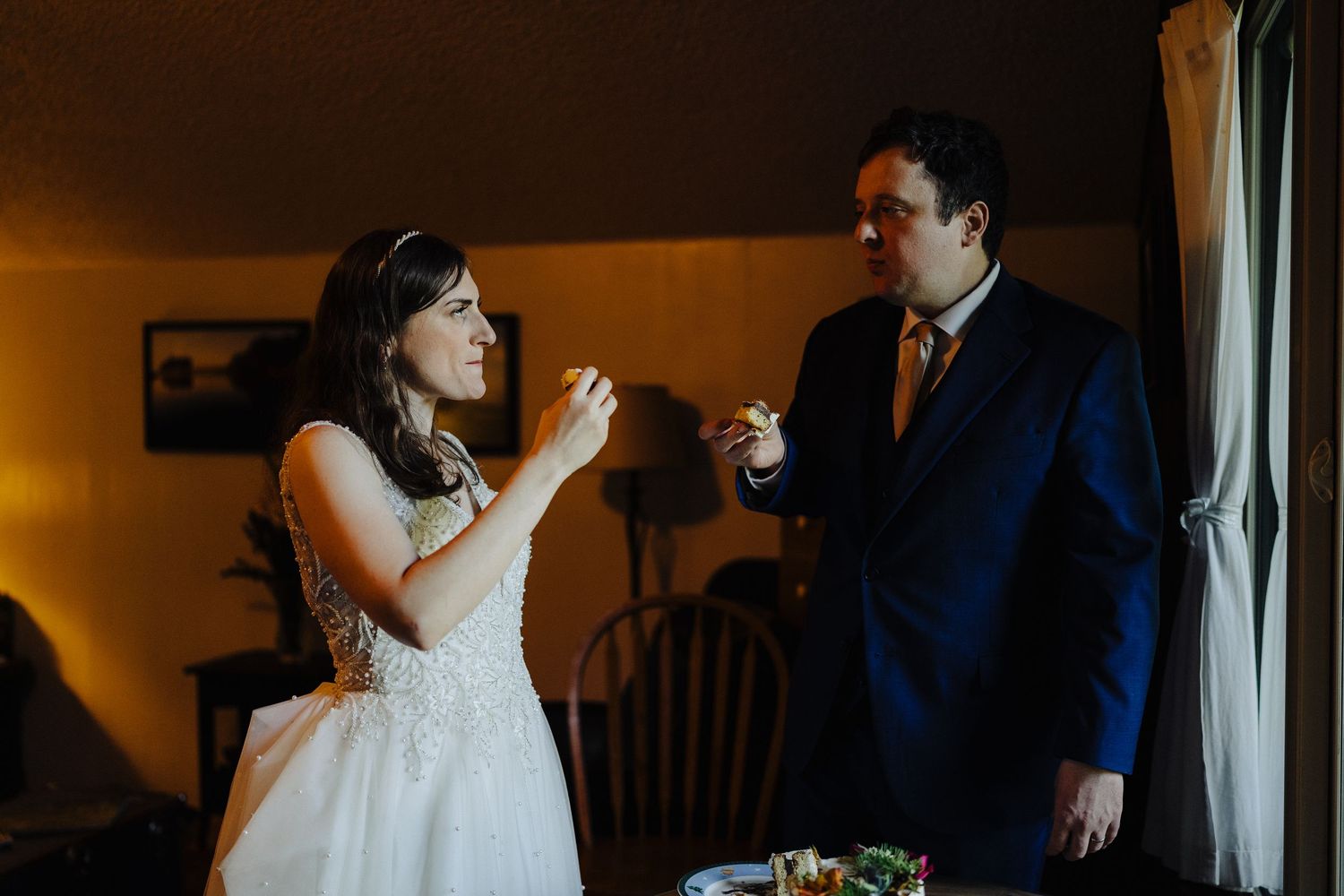 Two people in formalwear share cake together in a warmly lit room by a window.