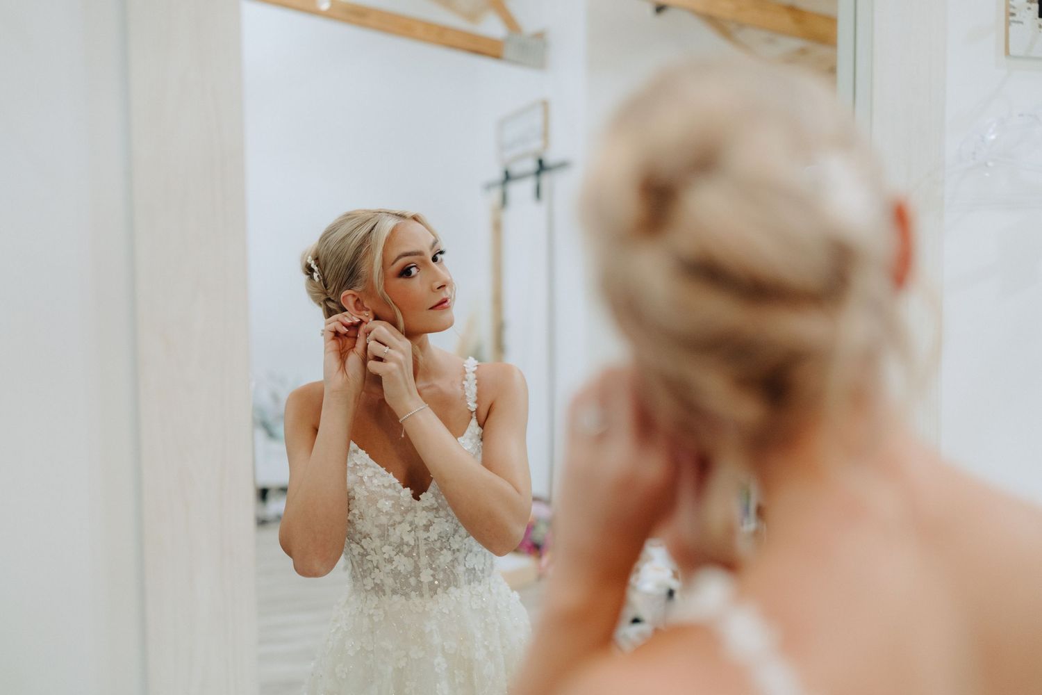 Woman in white dress adjusts her earrings while looking in a mirror during wedding preparations.