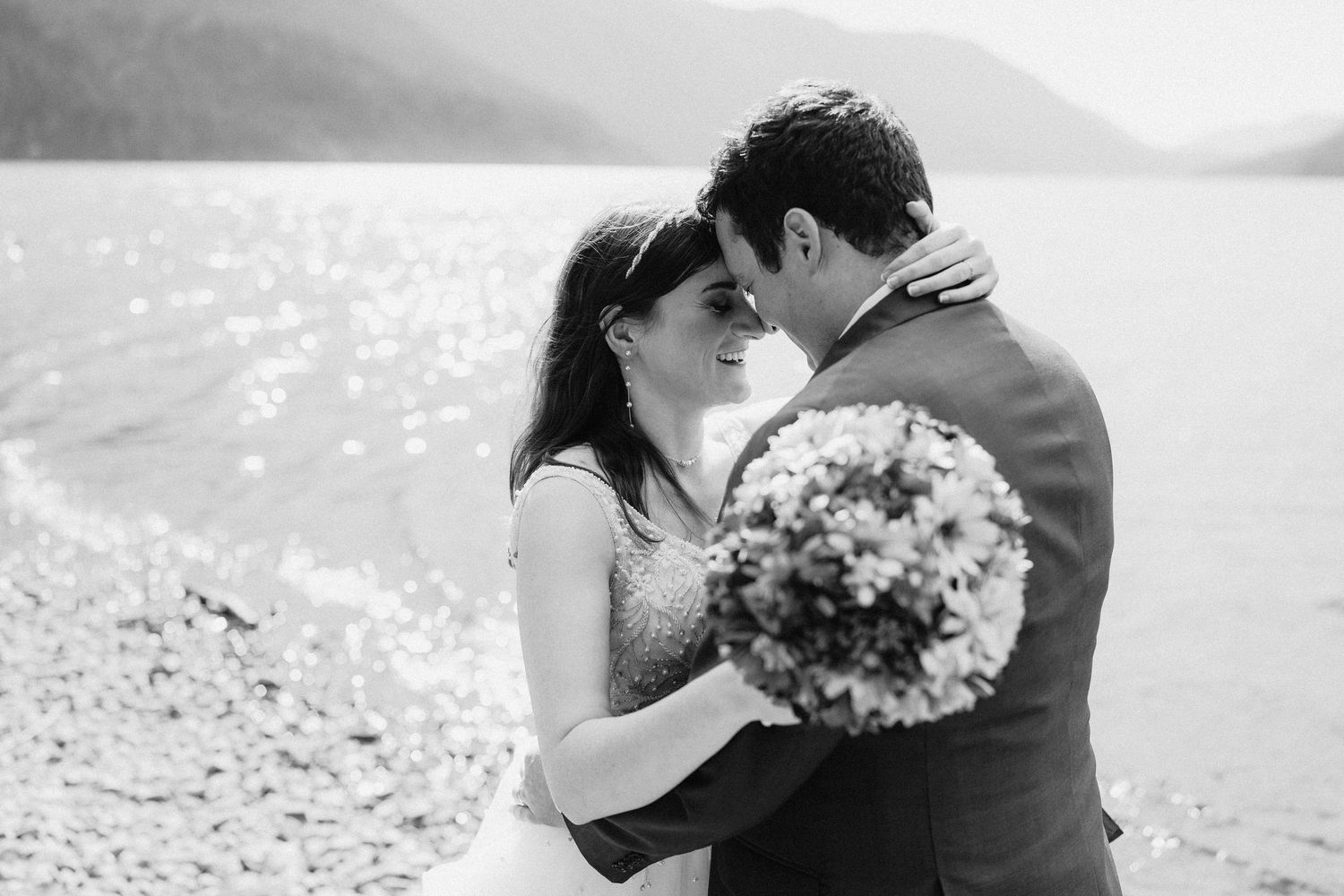 Black and white portrait of wedding couple sharing intimate moment by mountain lake.