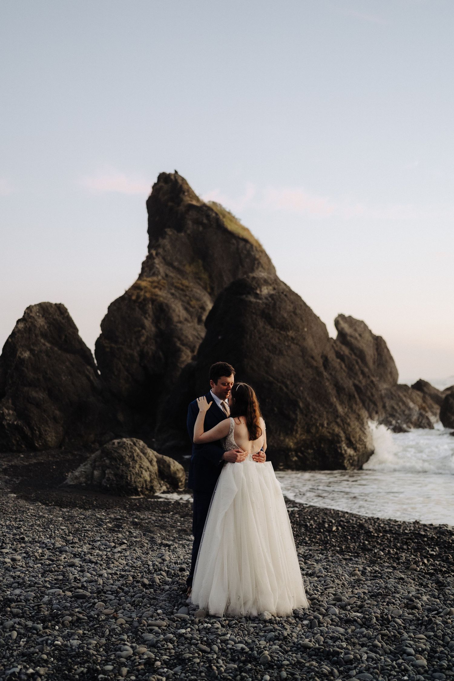 A couple in wedding attire embraces on a rocky beach with dramatic coastal rock formations at sunset.
