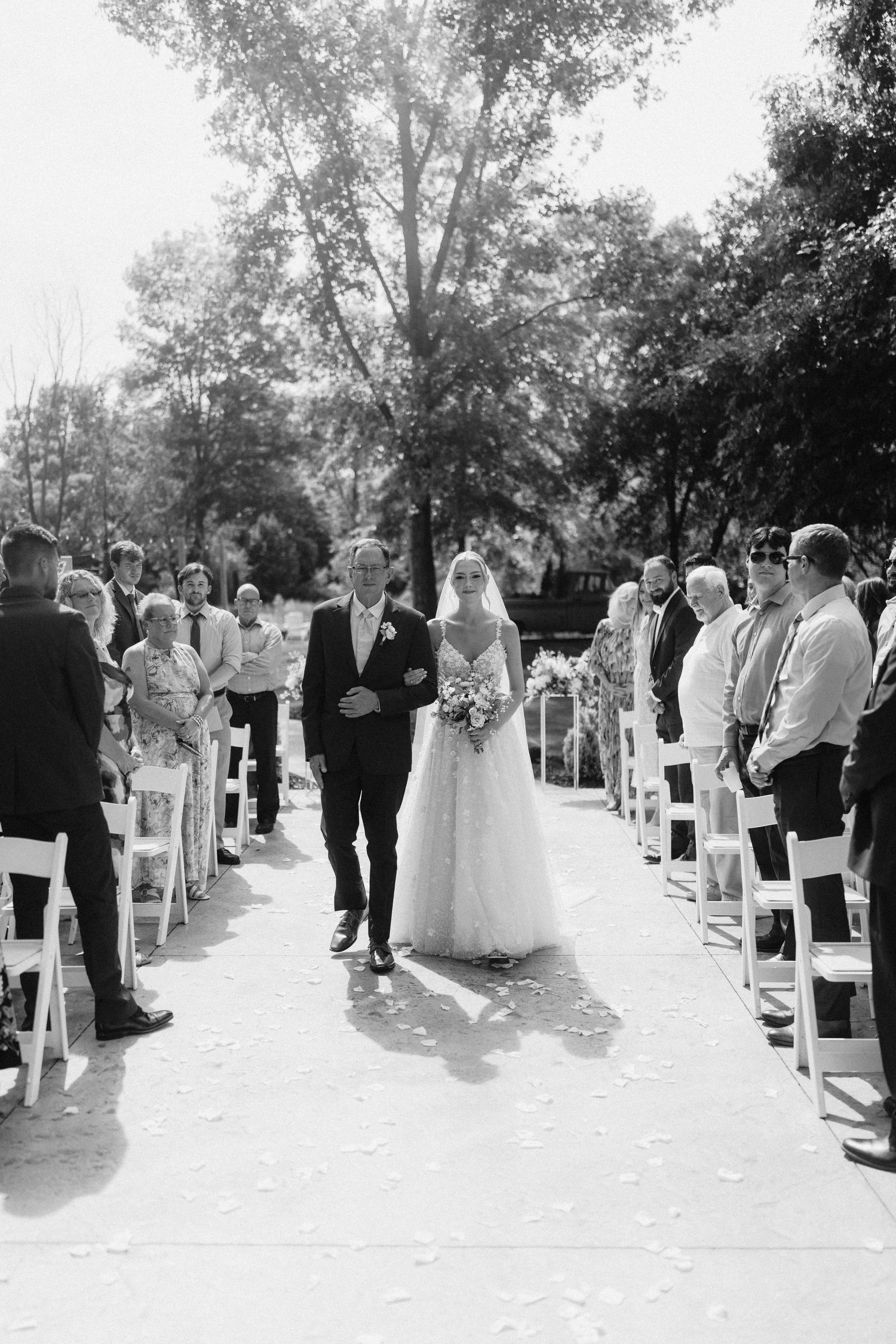 Black and white photo of wedding ceremony guests seated in white chairs along a tree-lined aisle.