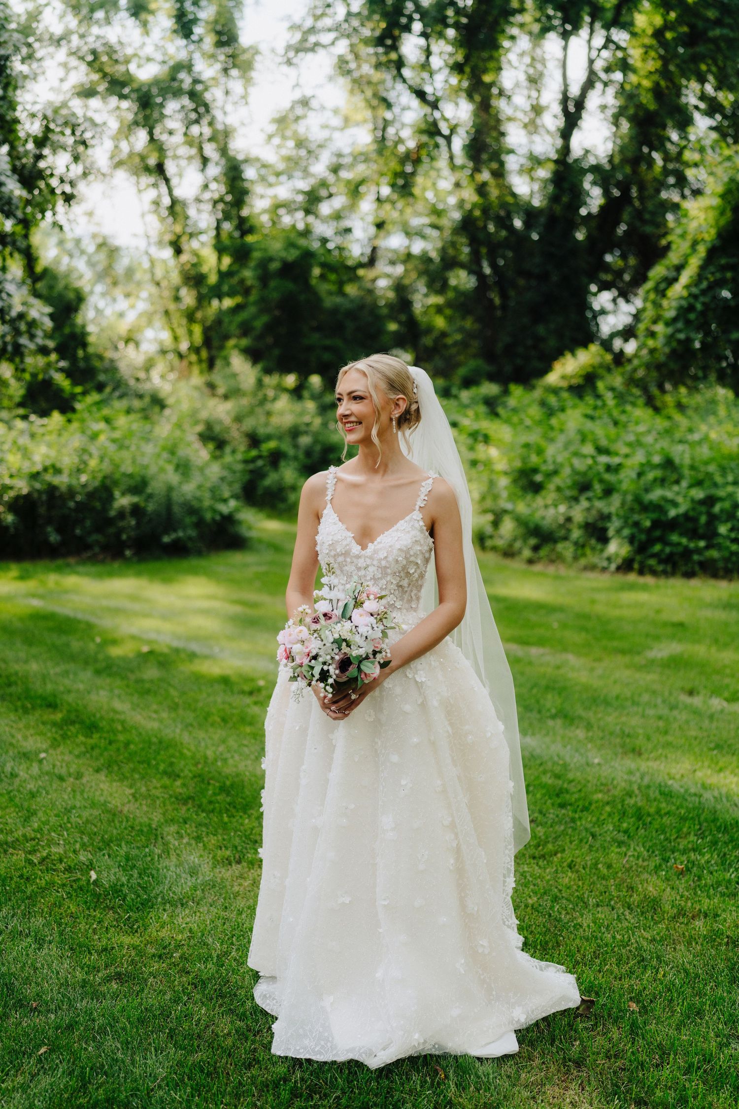 Bride in flowing white gown holding bouquet while standing on green grass.