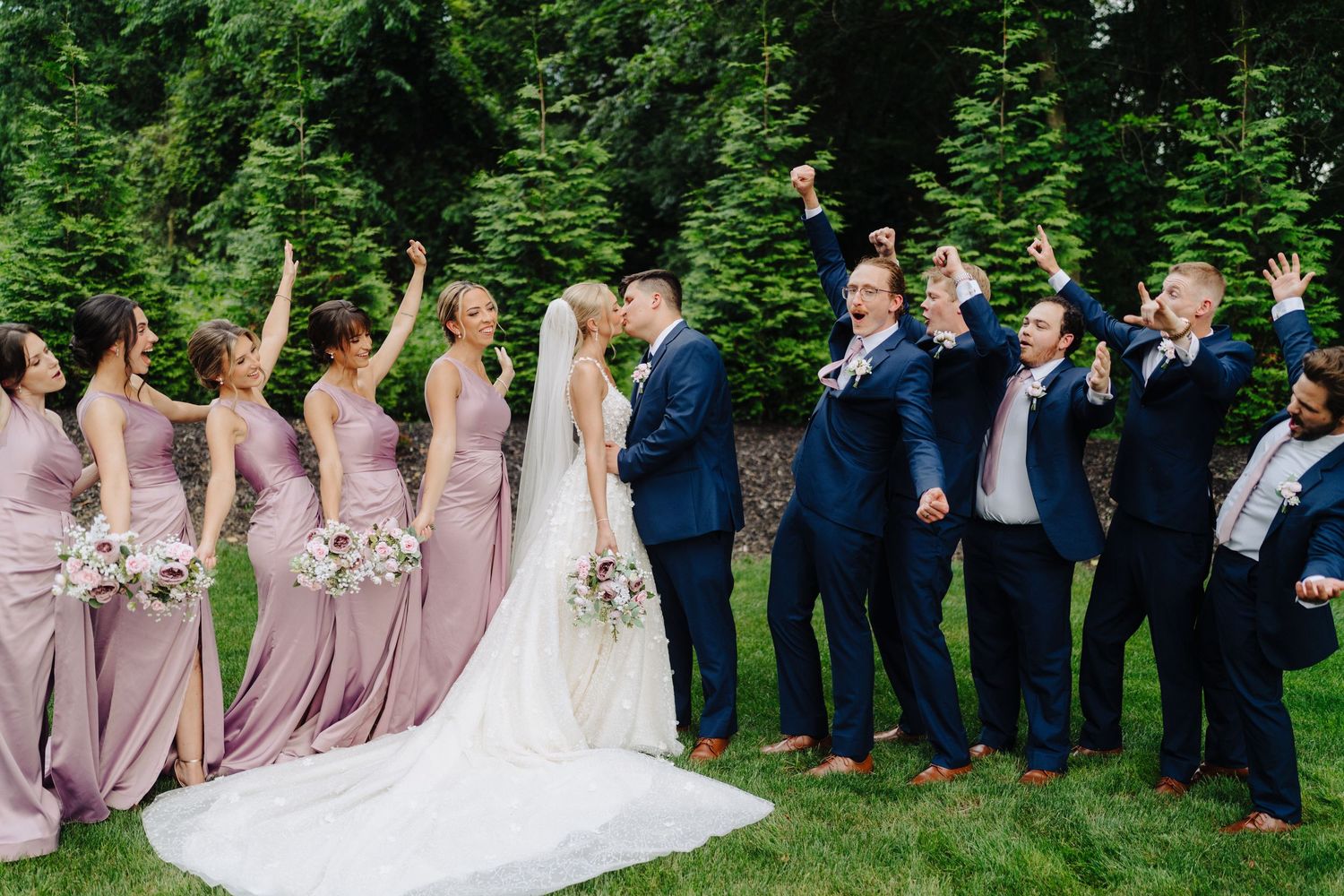 Wedding party in formal attire poses outdoors with bridesmaids in mauve dresses and groomsmen in navy suits.