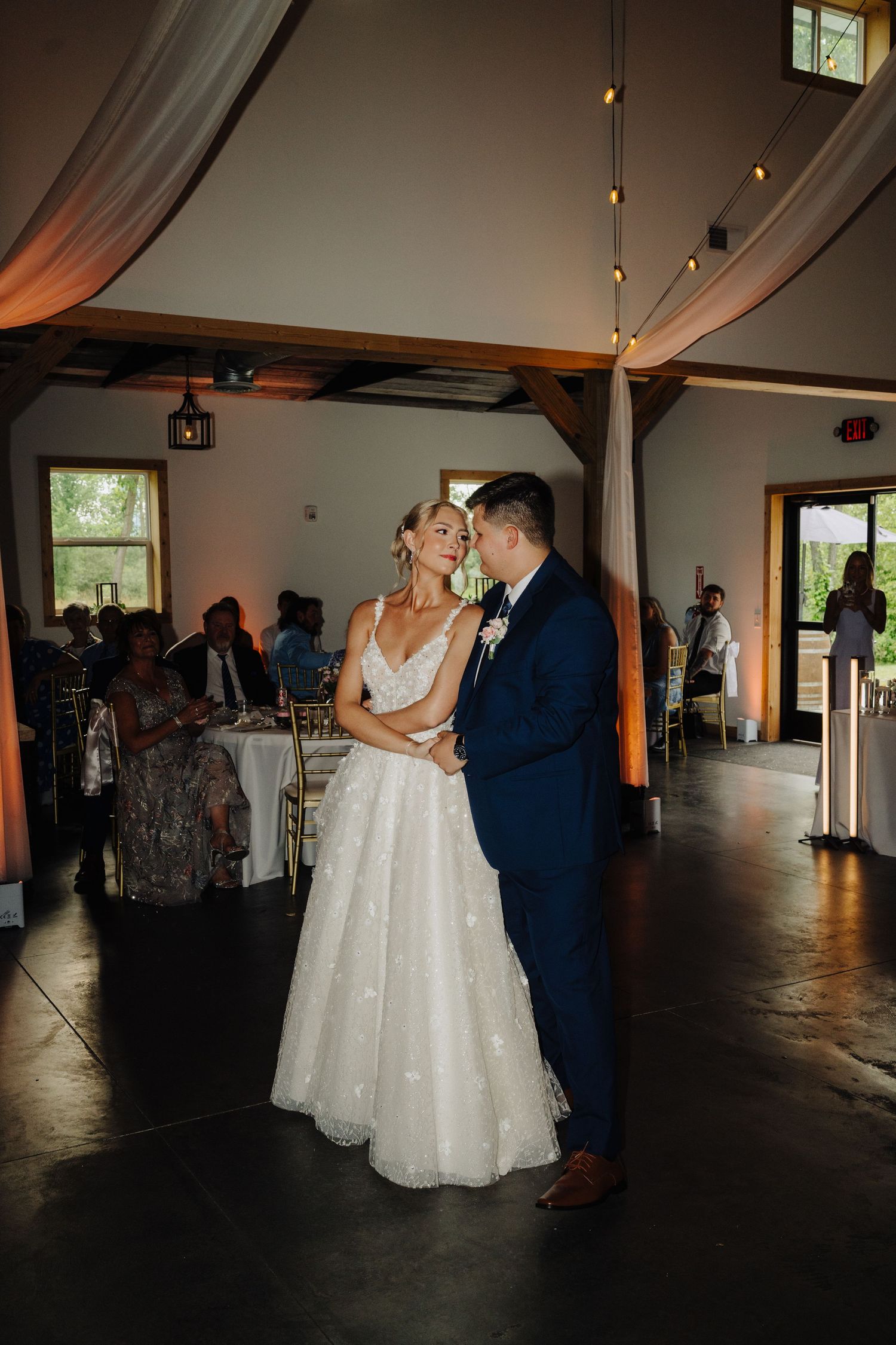 A romantic dance moment at a wedding reception in a rustic venue with draped fabric and warm lighting.