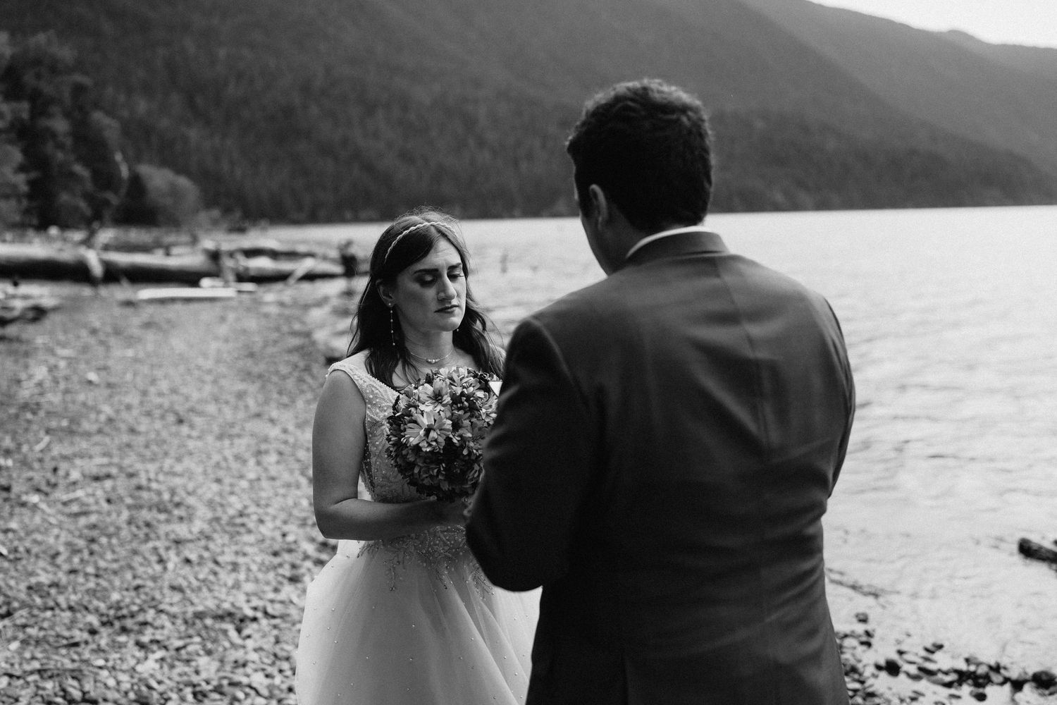 A couple shares a romantic moment on a rocky beach shore with mountains and water in the background, captured in black and white.