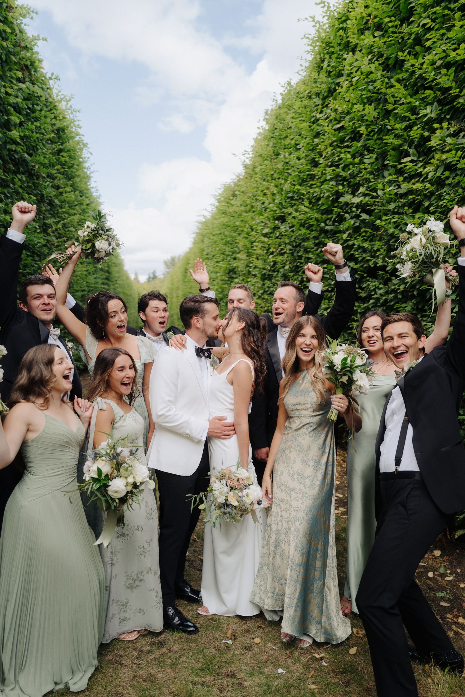 Wedding party celebrating outdoors with raised arms and joyful expressions among green hedges in garden setting.