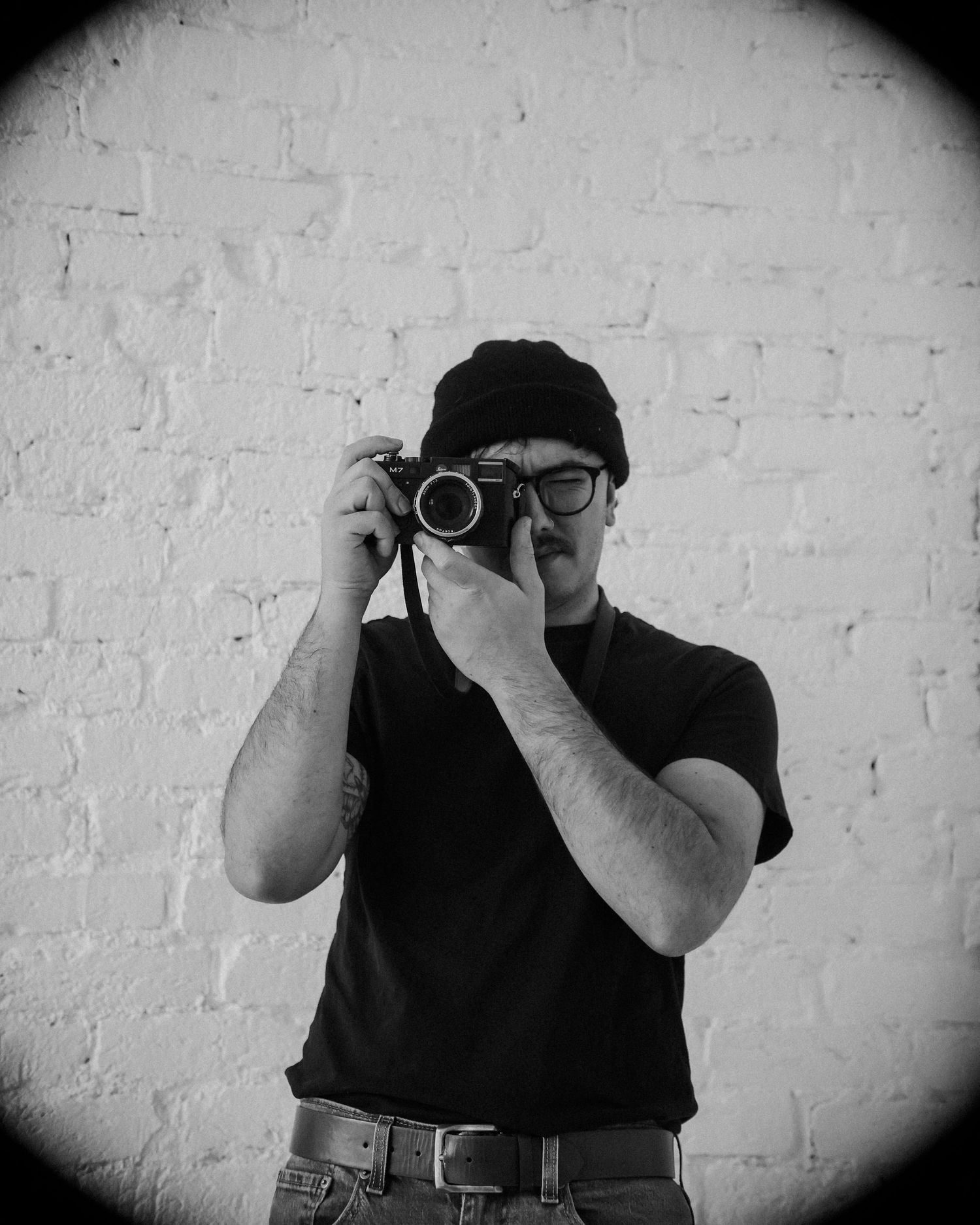 Black and white sequence of a photographer with a beanie and glasses taking photos against a white brick wall.