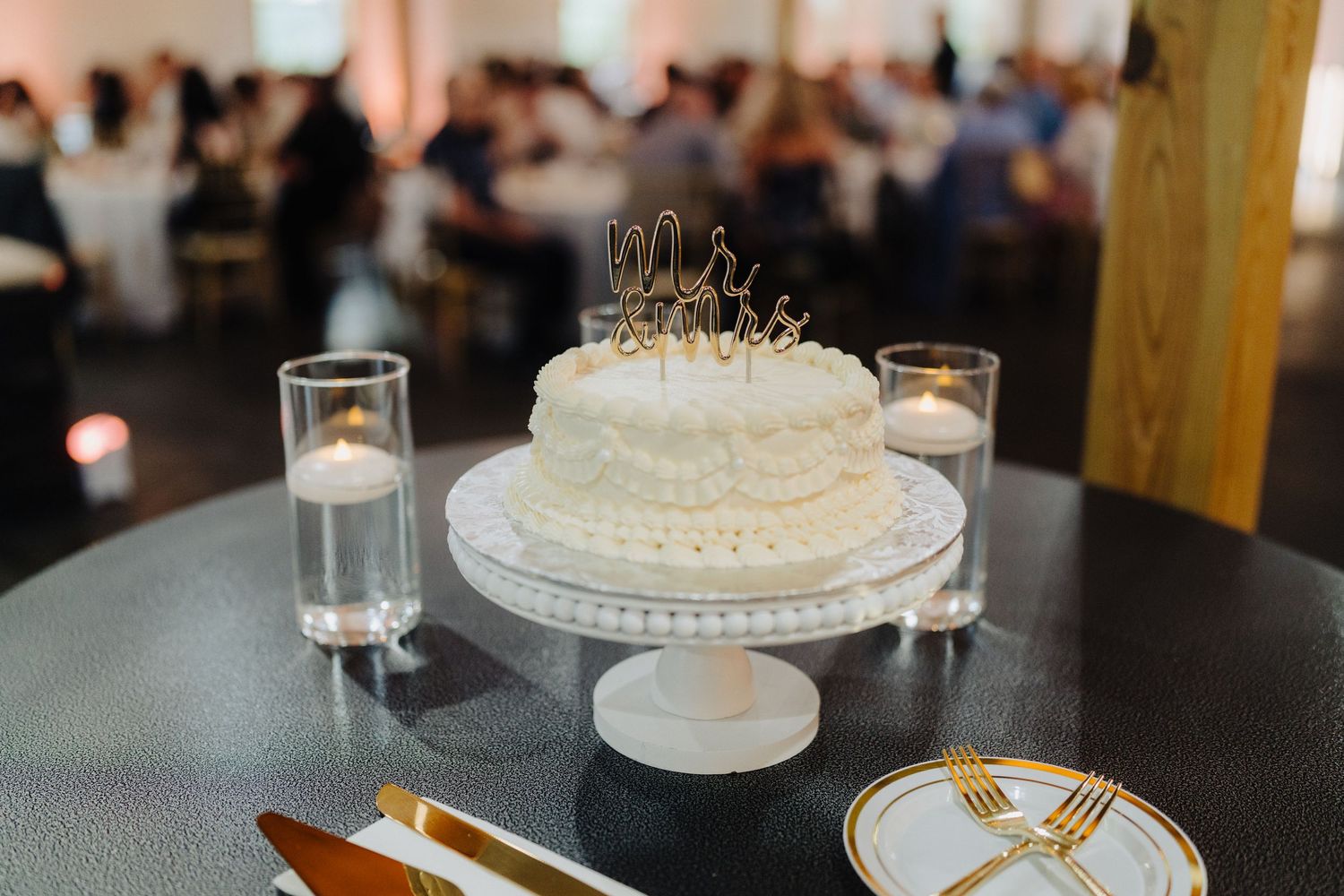 Elegant white wedding cake displayed on black stand with glassware and place settings.