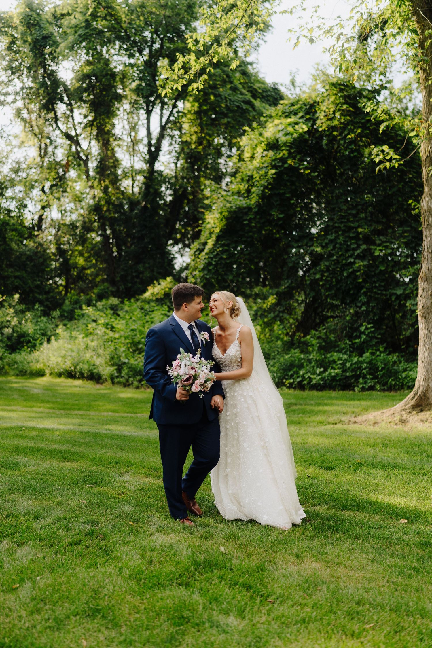 Newlyweds share an intimate moment on a sunlit lawn surrounded by trees.
