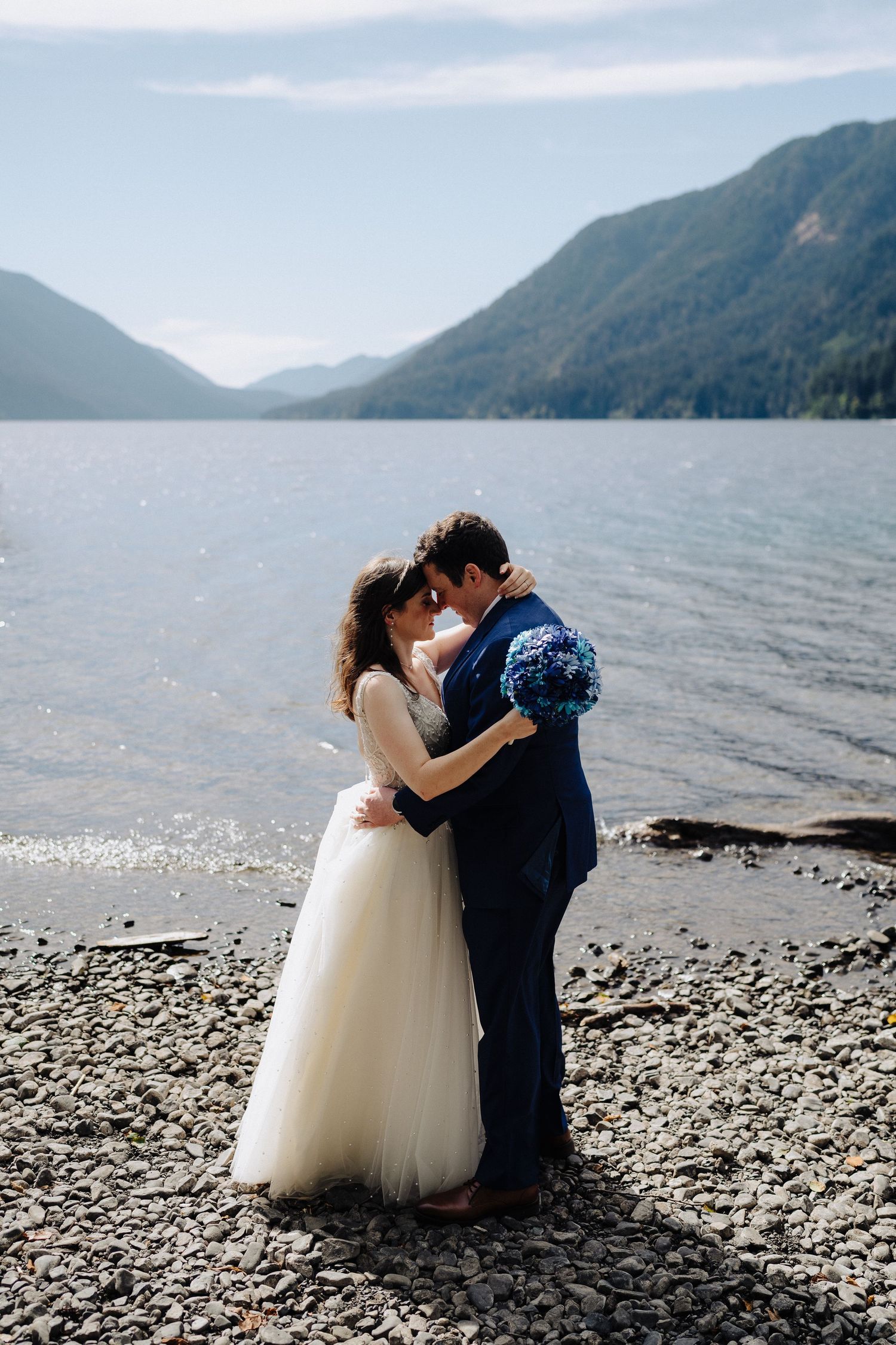 A romantic embrace between newlyweds on a pebble beach with mountains and lake in the background.