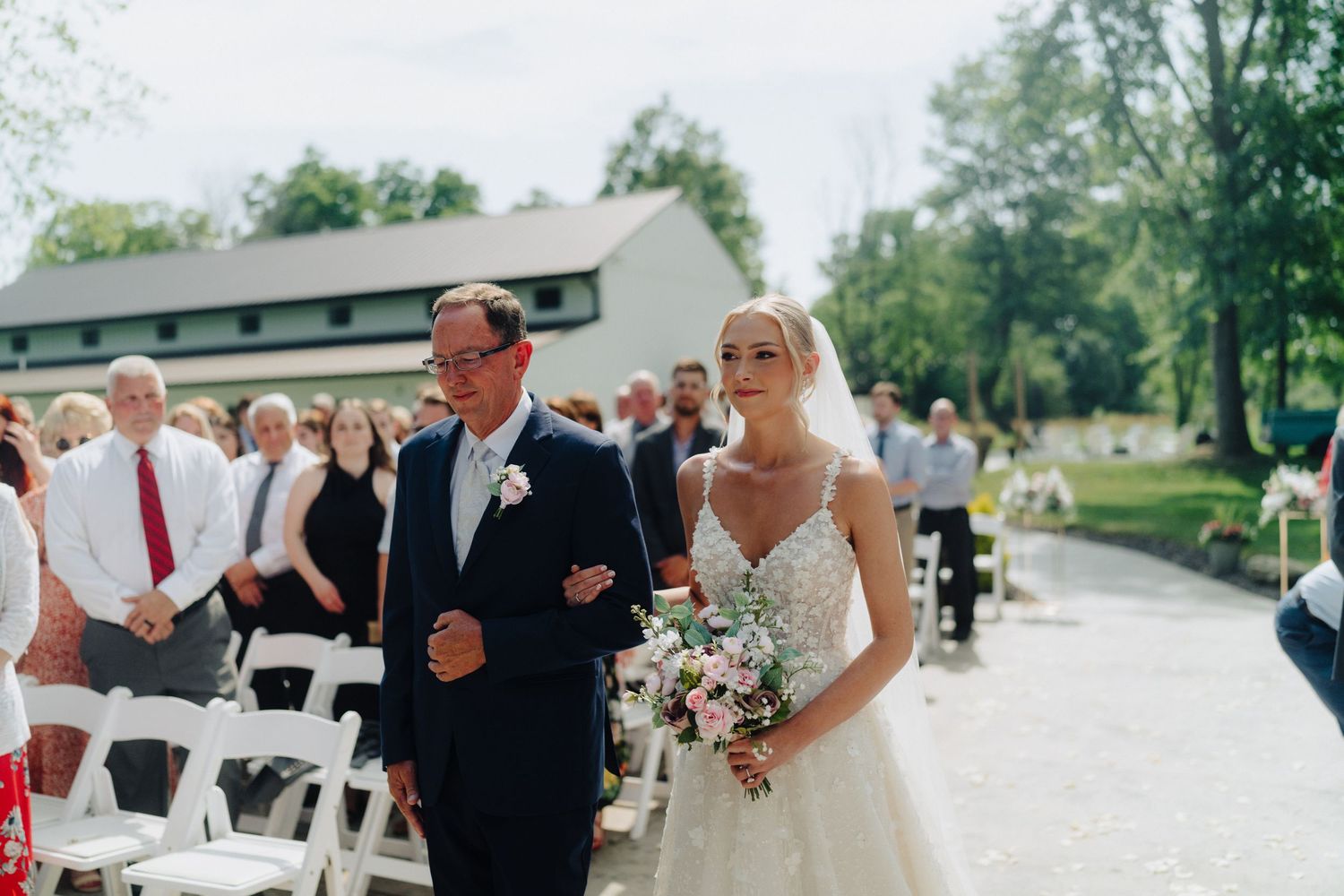 A wedding processional with a bride in white gown holding a bouquet walking down an outdoor aisle.
