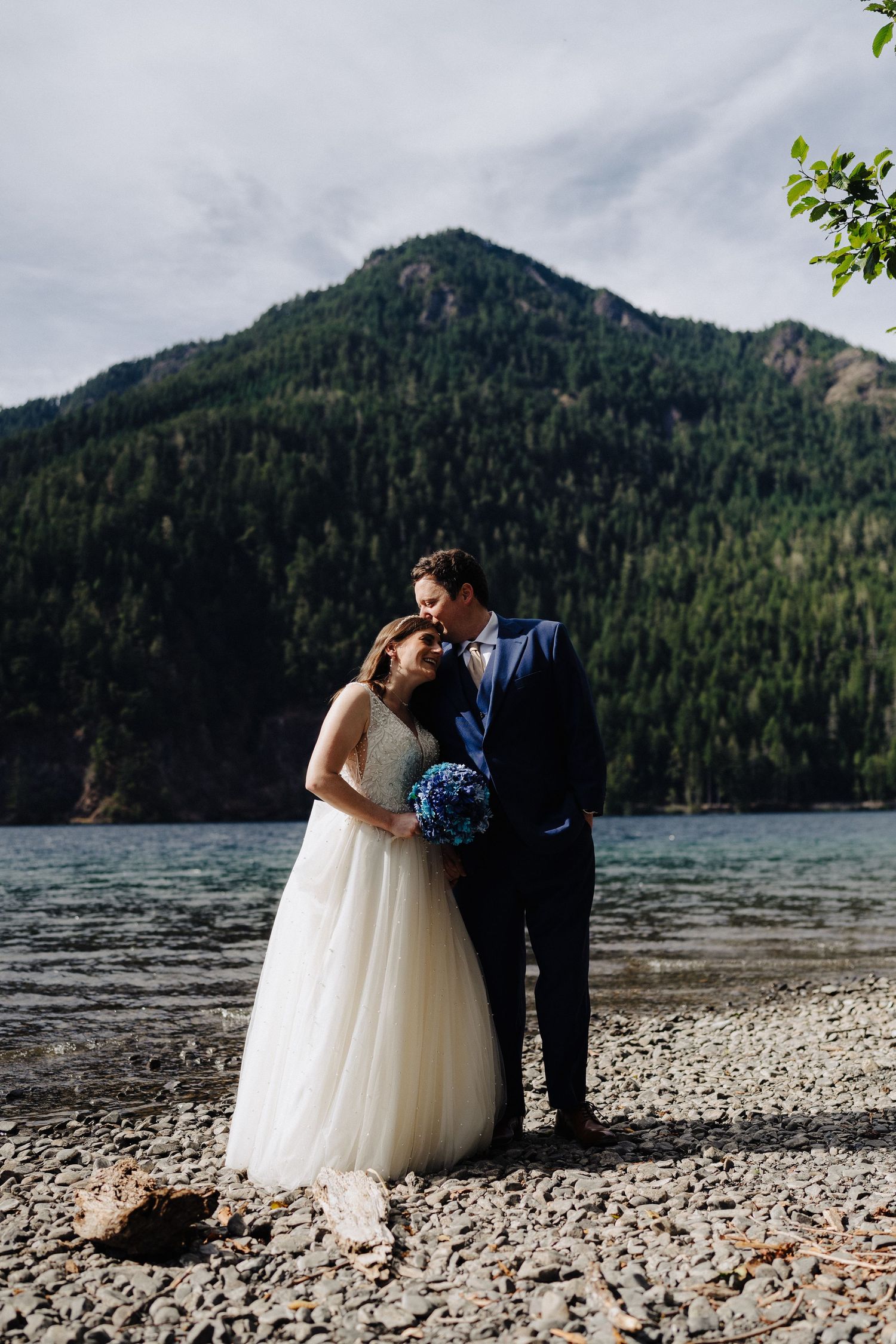 A romantic lakeside wedding photo with mountains and evergreen forest in the background.