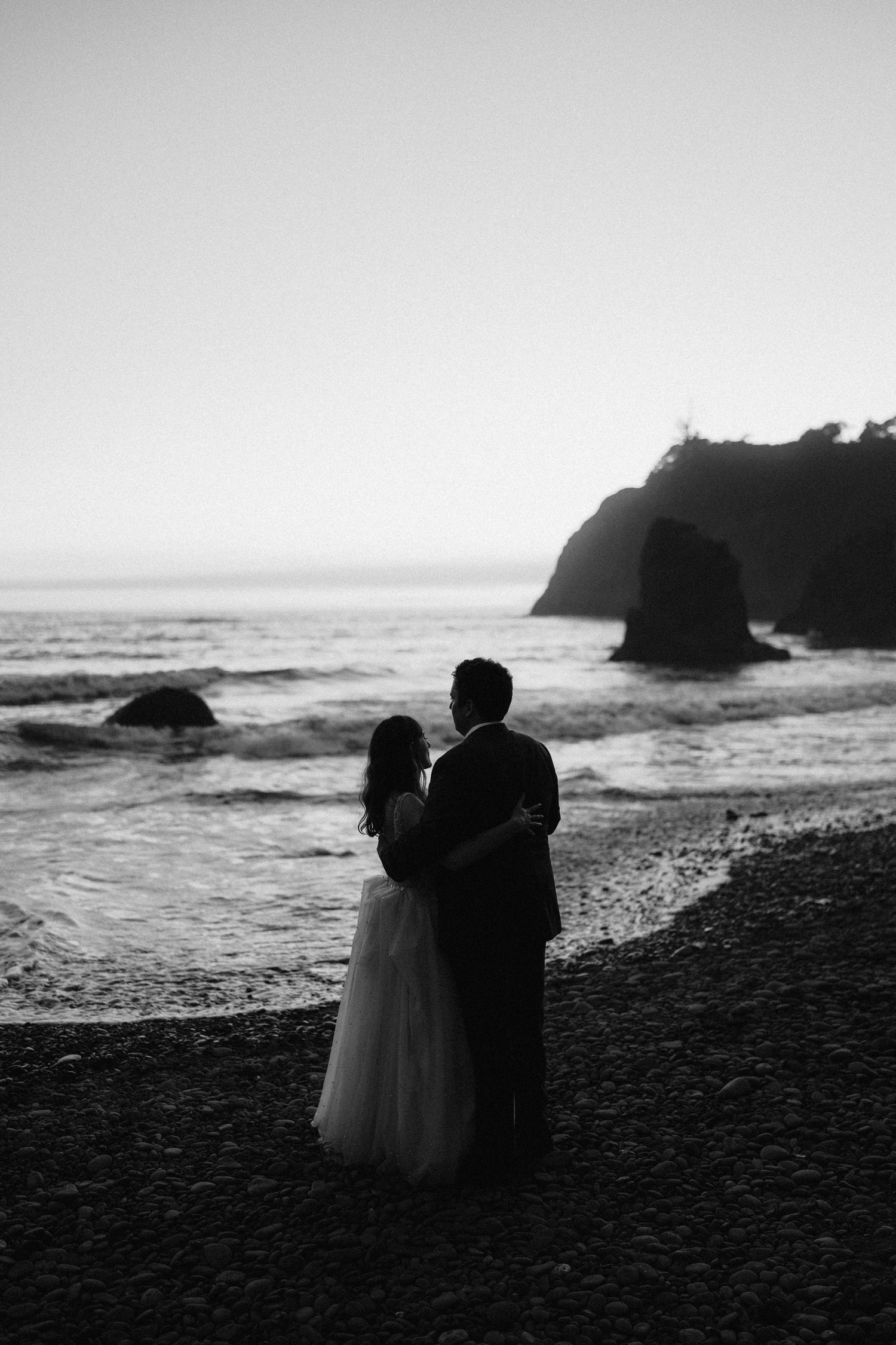 A couple in wedding attire embraces on a rocky beach with dramatic cliffs in black and white.