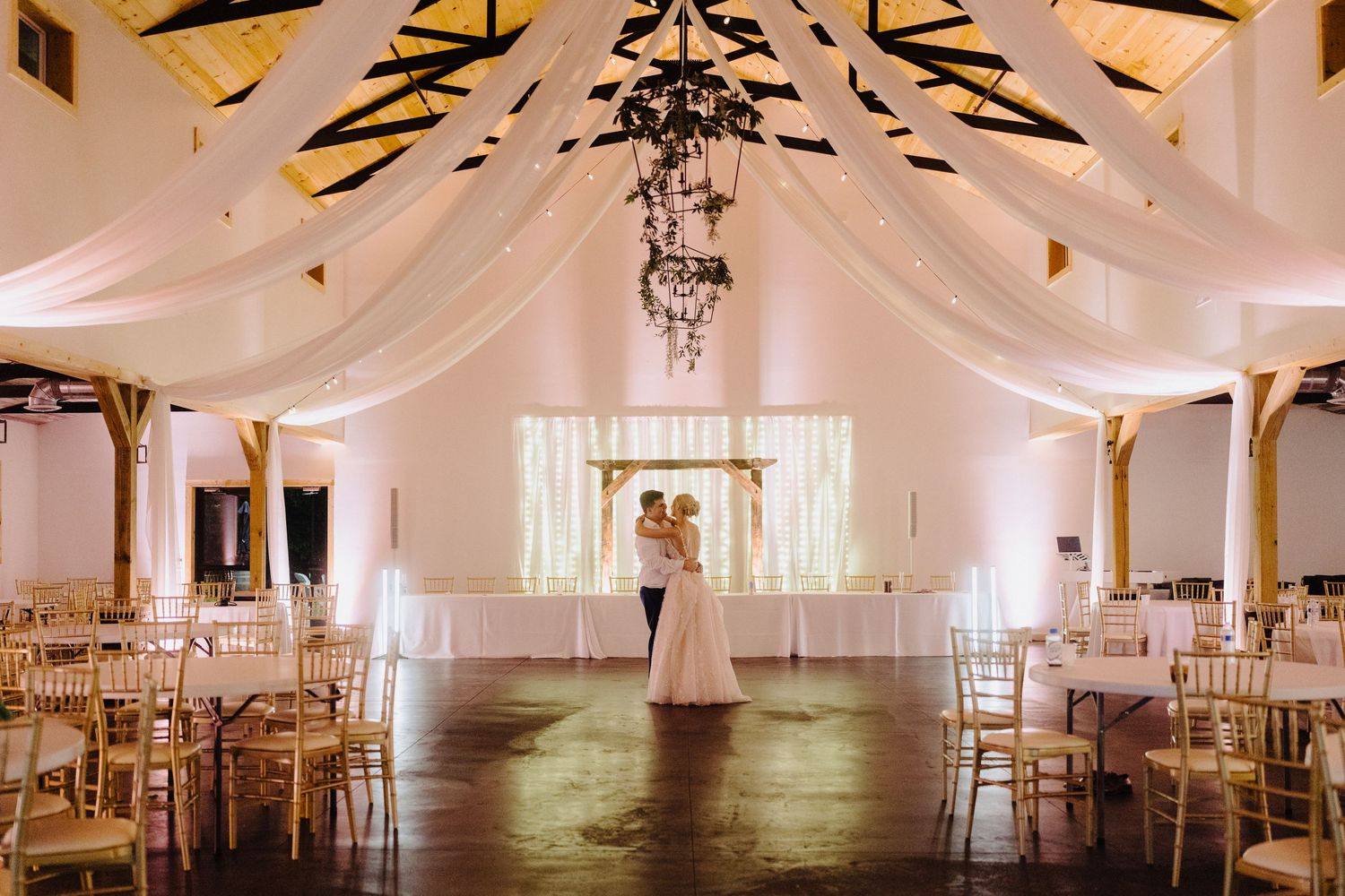 Elegant white wedding reception hall with draped fabric and golden chiavari chairs.