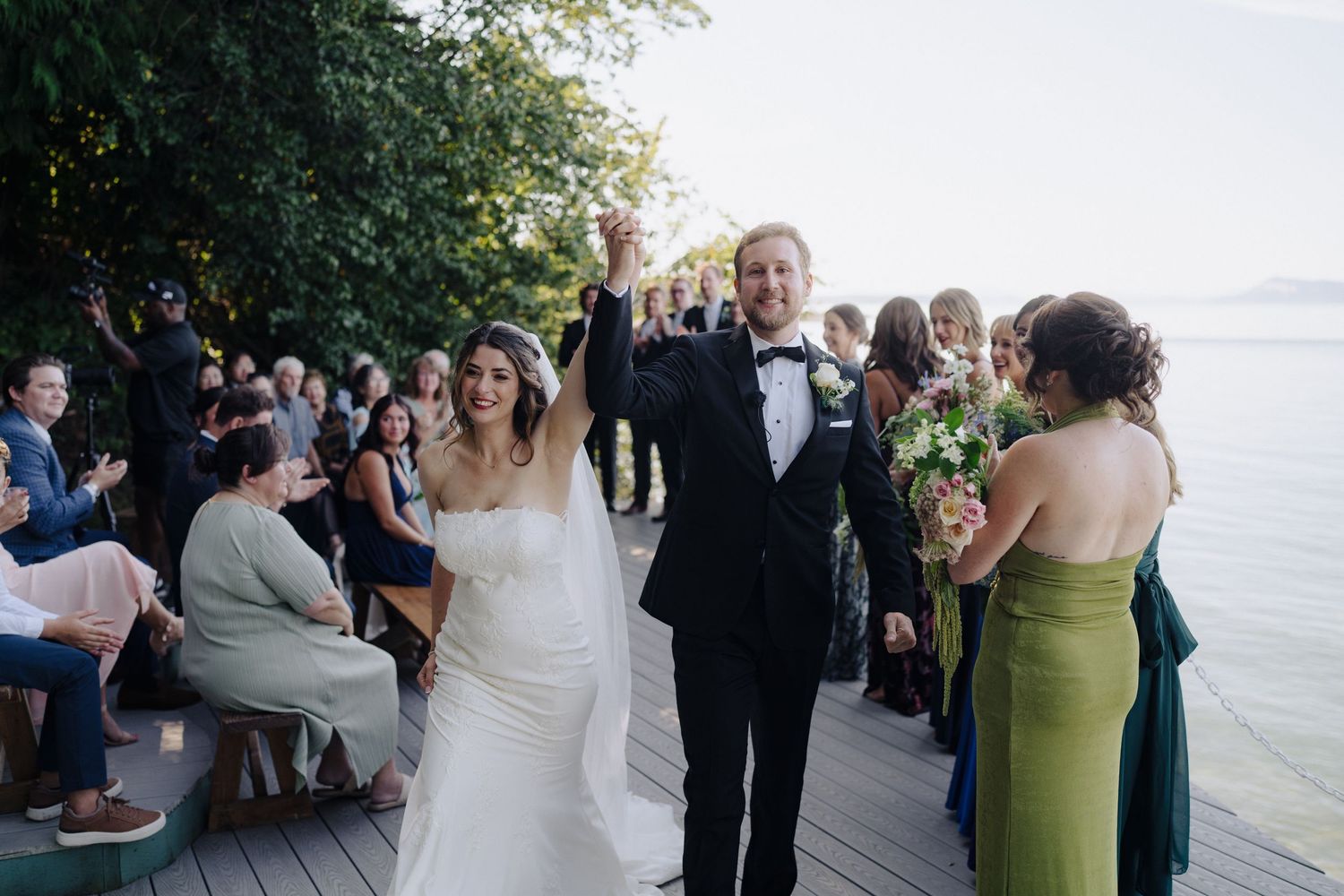 Newlyweds walk down outdoor deck by waterfront during sunset celebration with guests looking on.
