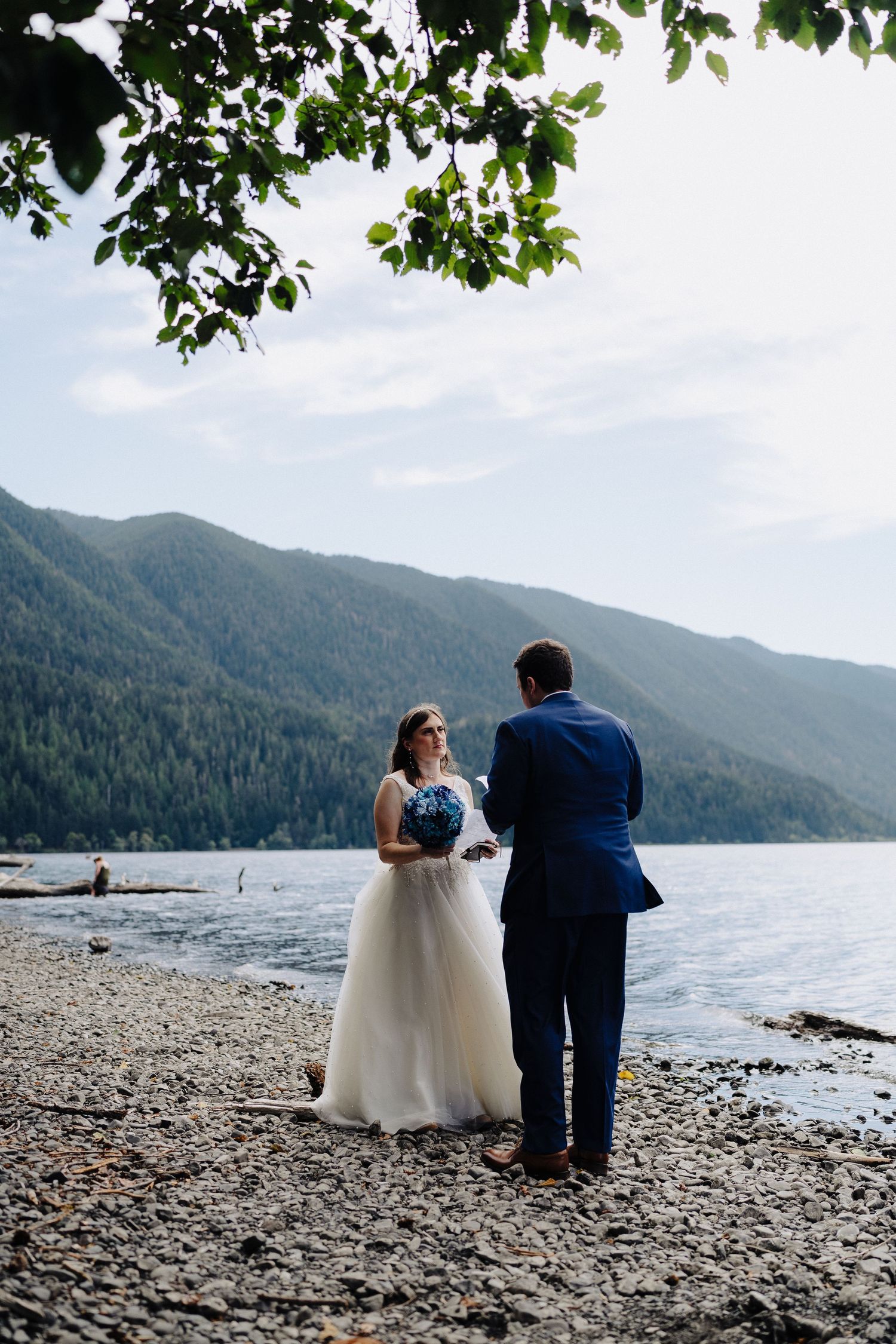 Newlyweds share a moment on a serene lakeside beach with mountains in the background.