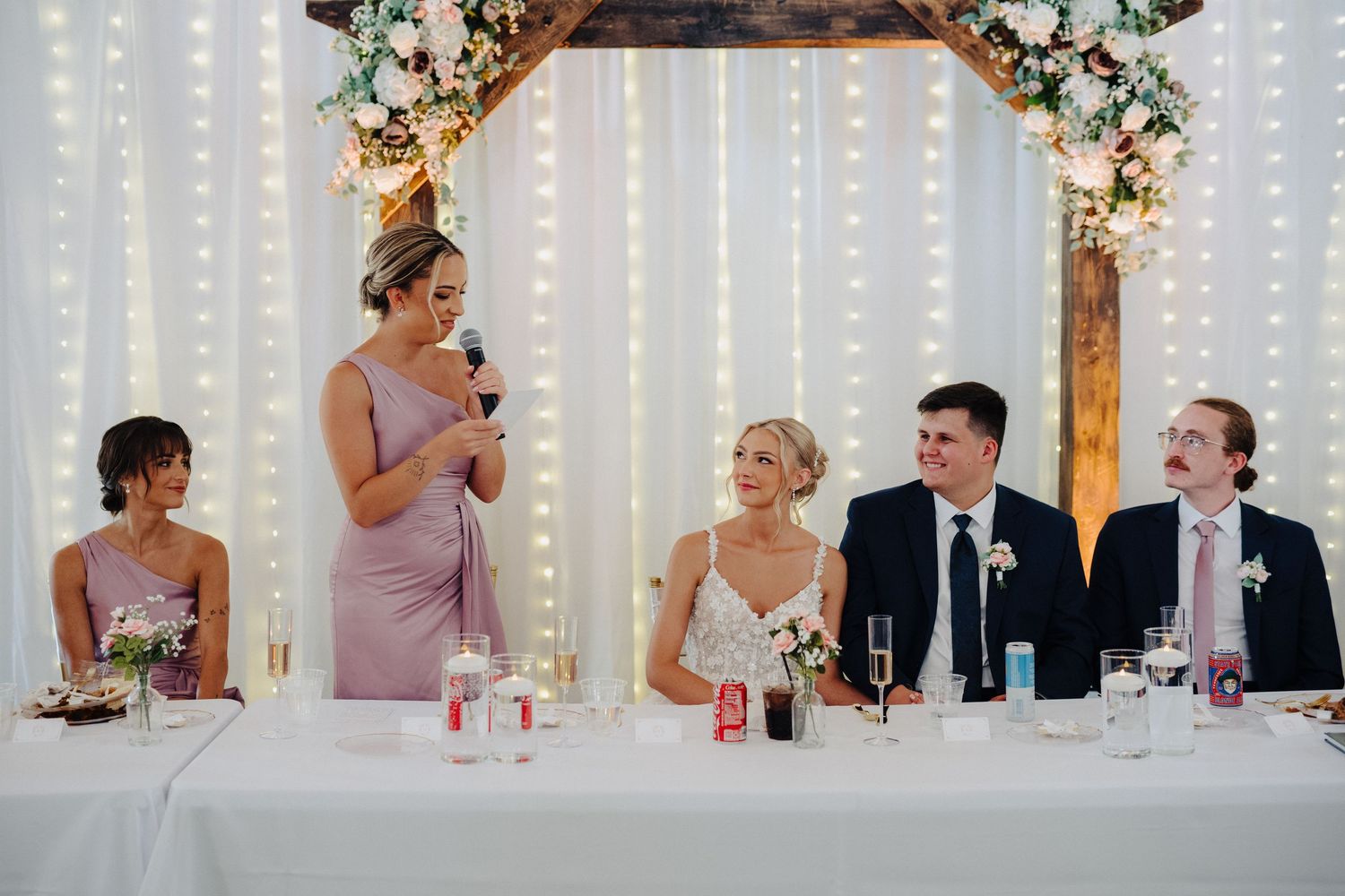 Wedding reception with decorated arch, string lights, and head table during speeches.