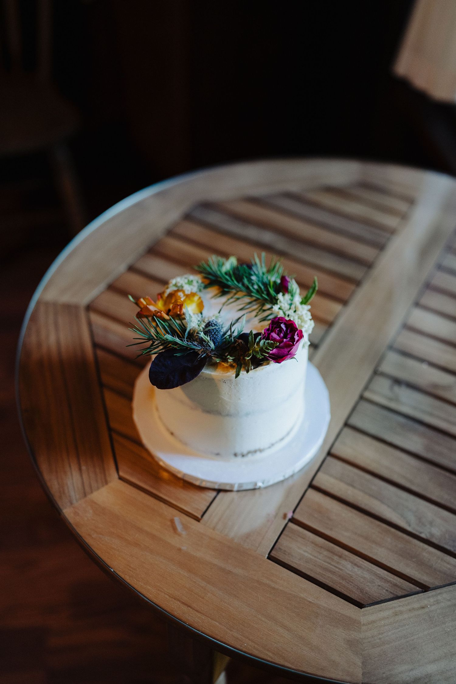 Small white wedding cake decorated with colorful wildflowers and greenery sits on a wooden round table.