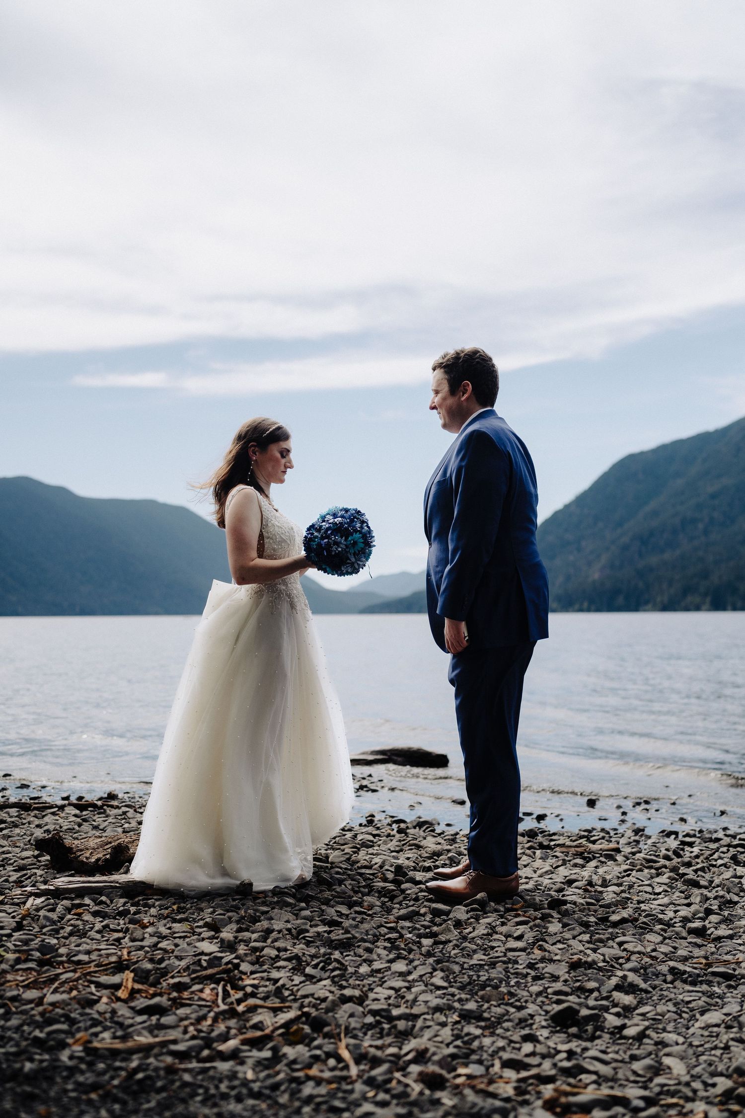 A couple in wedding attire stands on a rocky beach beside a lake surrounded by mountains.