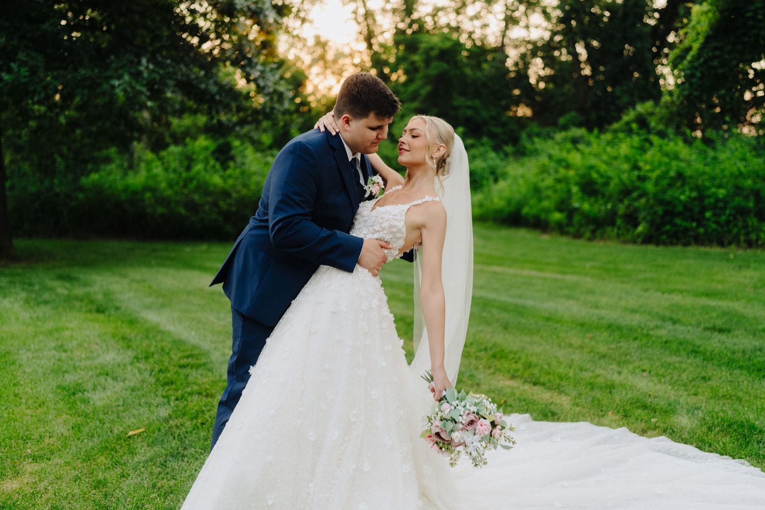 Newlyweds share romantic moment on green lawn during golden hour.