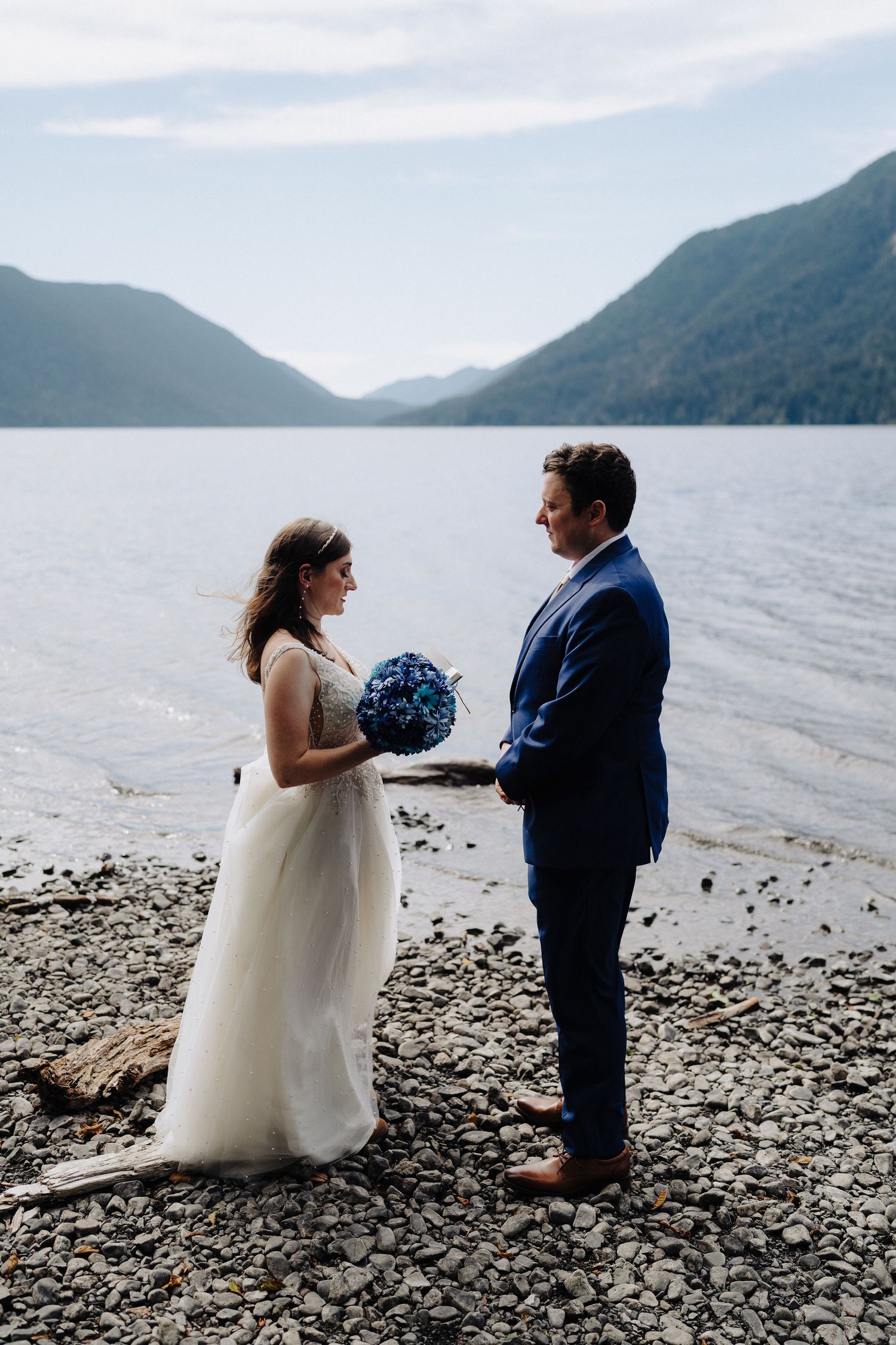 A newlywed couple stands on a rocky beach holding a blue bouquet with mountains and lake in the background.