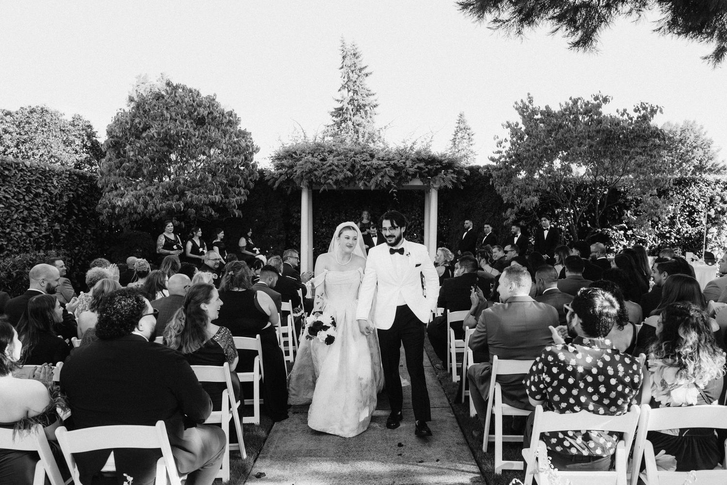 A black and white outdoor wedding ceremony shows a couple celebrating at the altar surrounded by seated guests in a garden setting.
