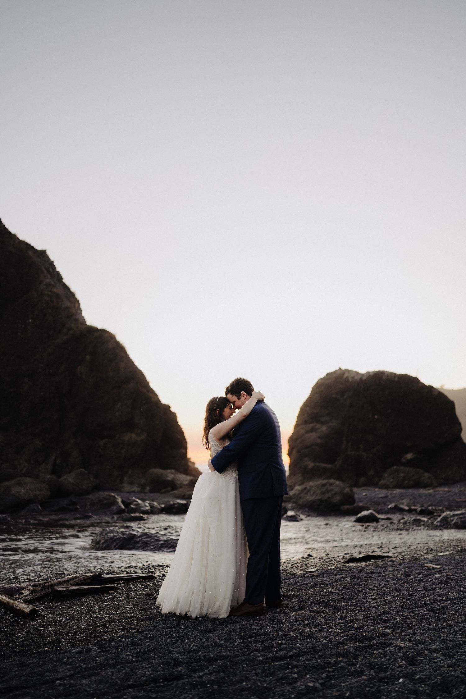 A couple embraces on a moody beach at sunset, surrounded by dramatic coastal rock formations.