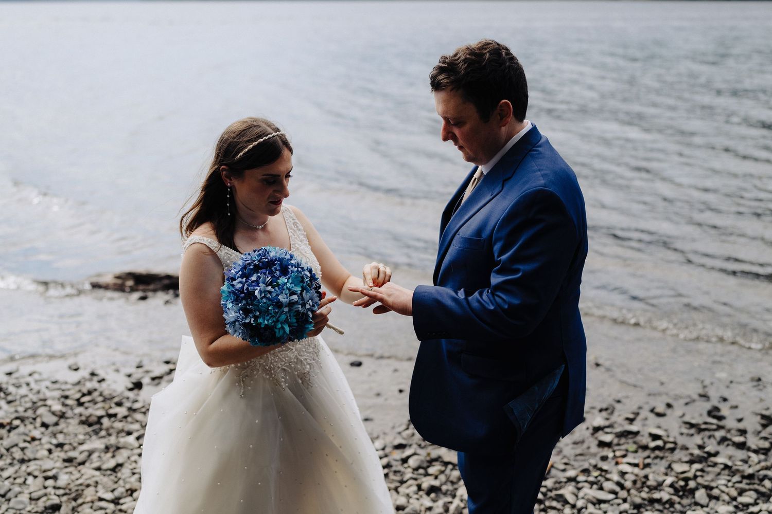 Wedding ceremony moment on rocky beach with mountains and lake in background.