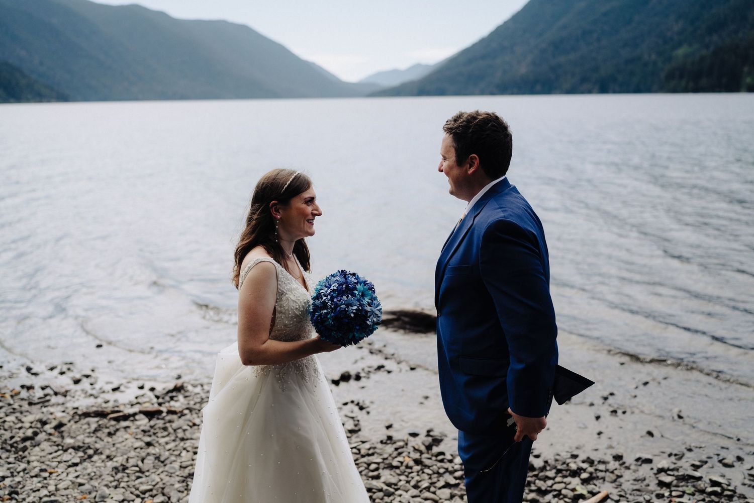 A couple shares a moment by a mountain lake, with the bride holding a blue bouquet.