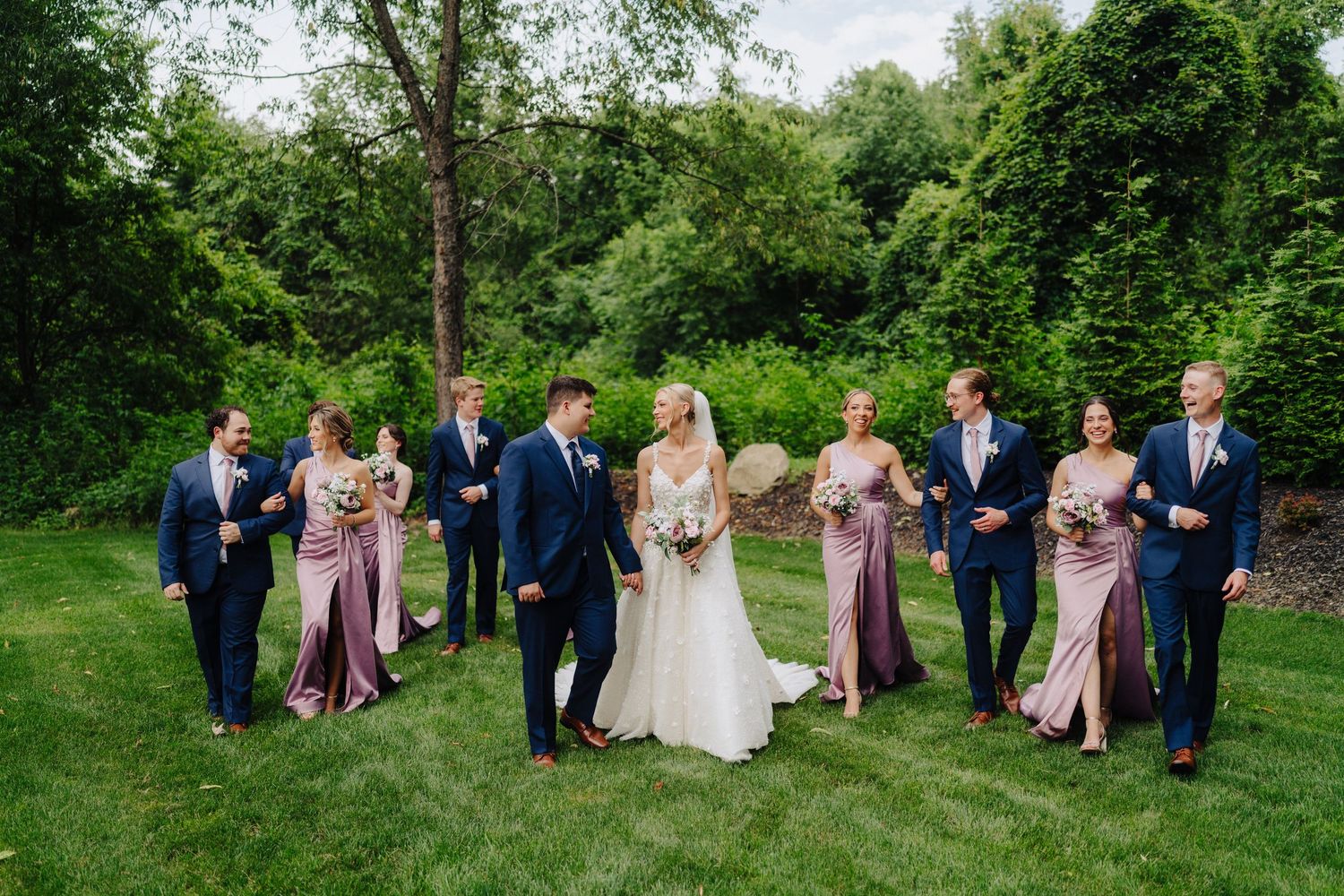 Wedding party in navy suits and mauve dresses poses together on lush green lawn.