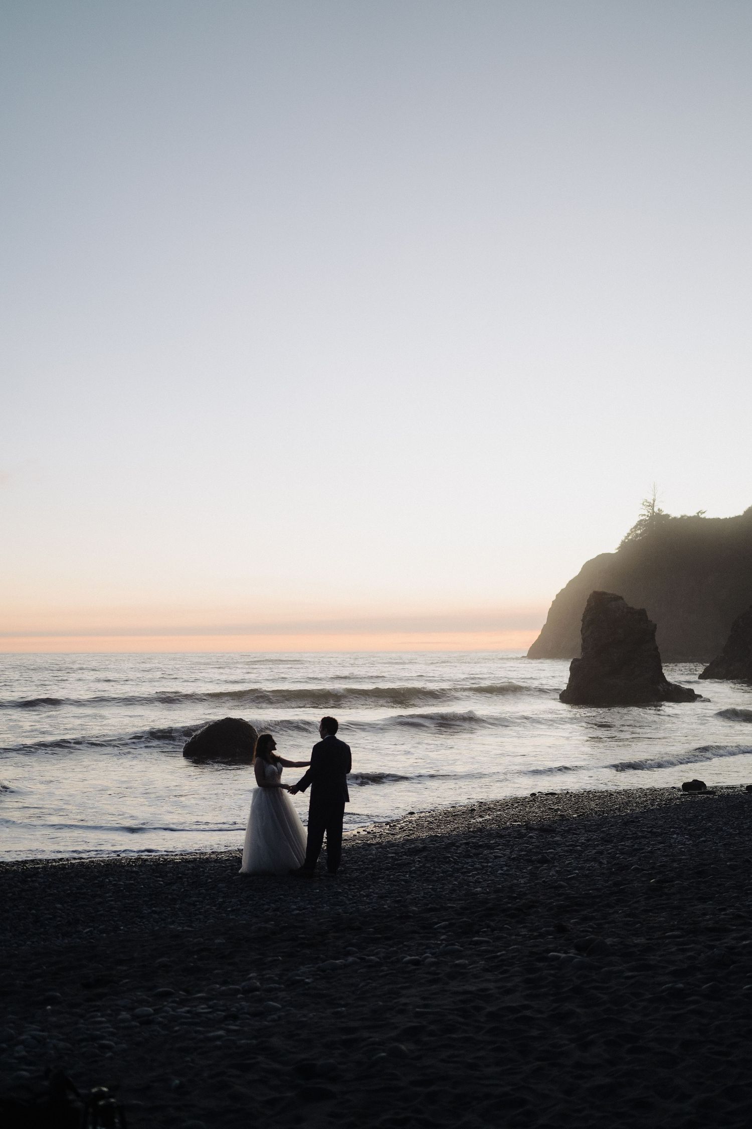 Silhouette of newlyweds on beach at dusk with lighthouse cliff in background.