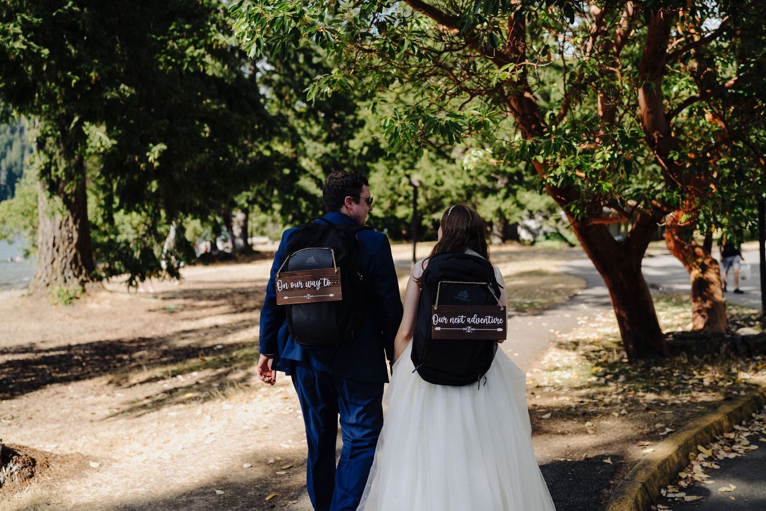 A couple walks down a tree-lined path wearing coordinated denim jackets over wedding attire.
