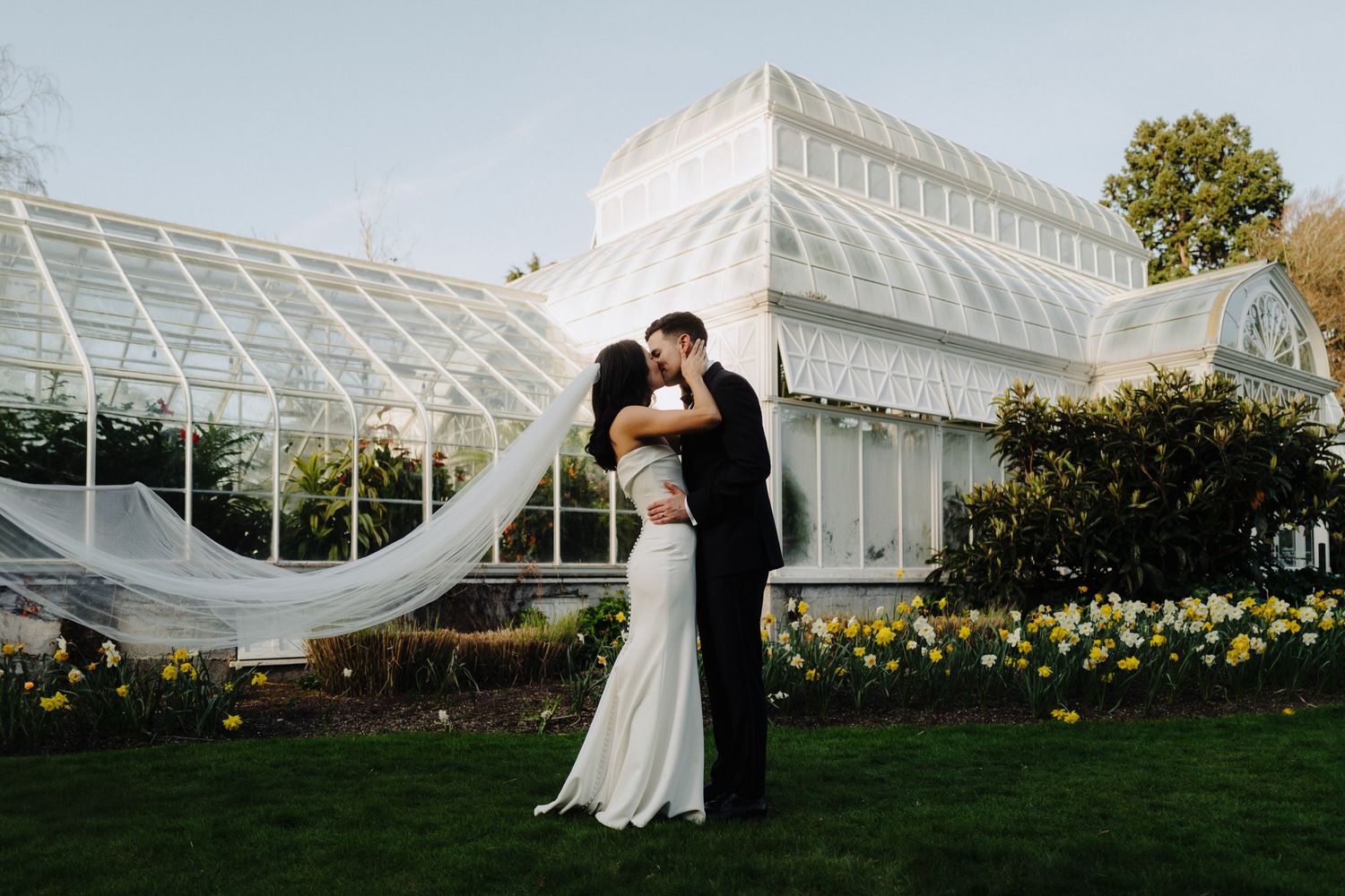 Couple embracing outside elegant glass conservatory with flowing white veil and garden surroundings.