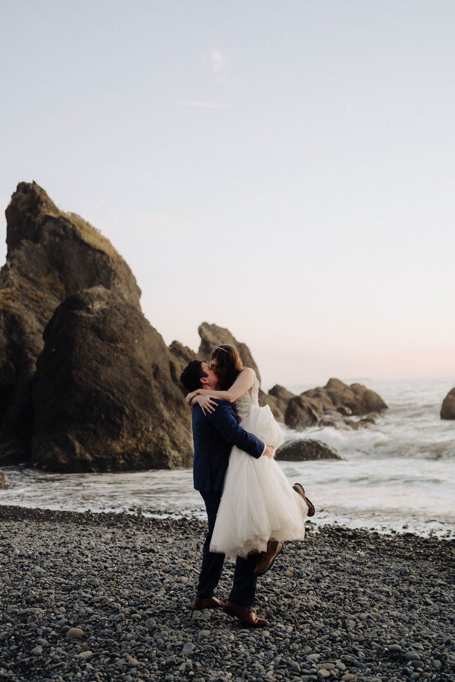Newlyweds embrace on rocky beach at sunset with dramatic ocean cliffs.