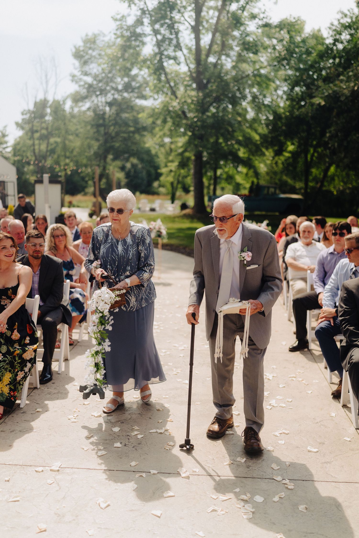 Elderly couple walking down outdoor ceremony aisle lined with seated guests.